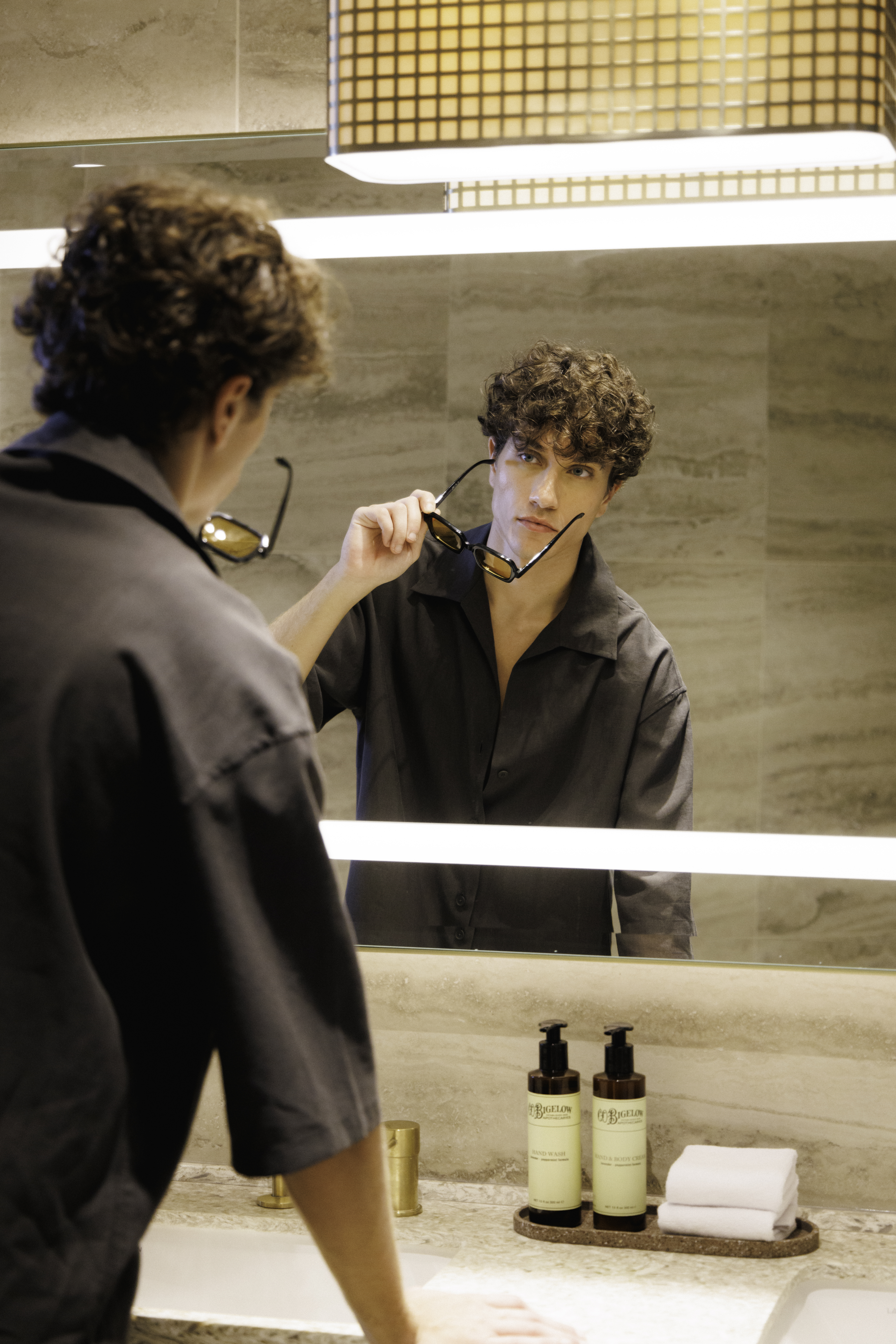Curly-haired man holds sunglasses, reflected in modern bathroom mirror.