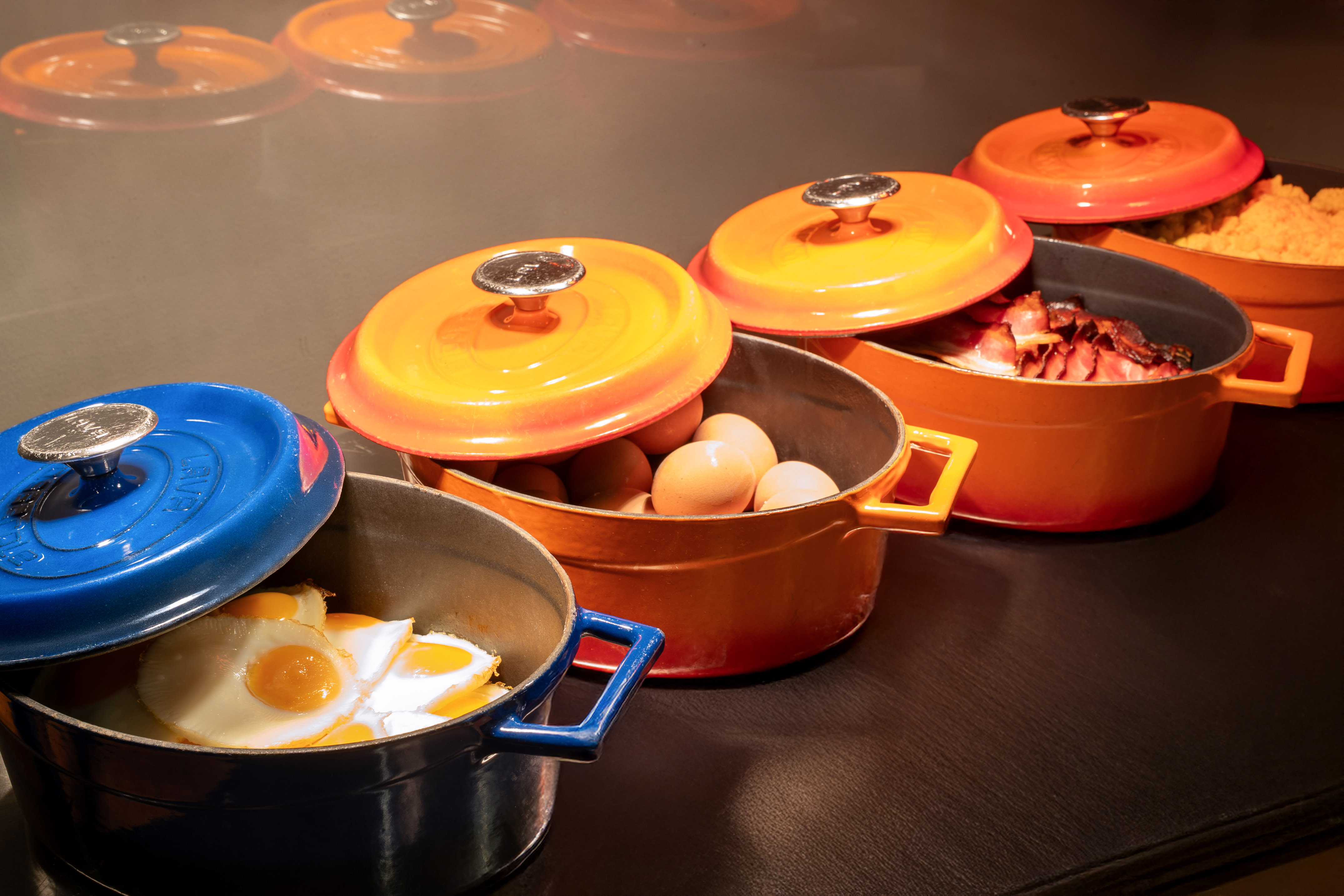 Breakfast buffet with colorful cast iron pots: fried eggs, bacon, raw eggs.