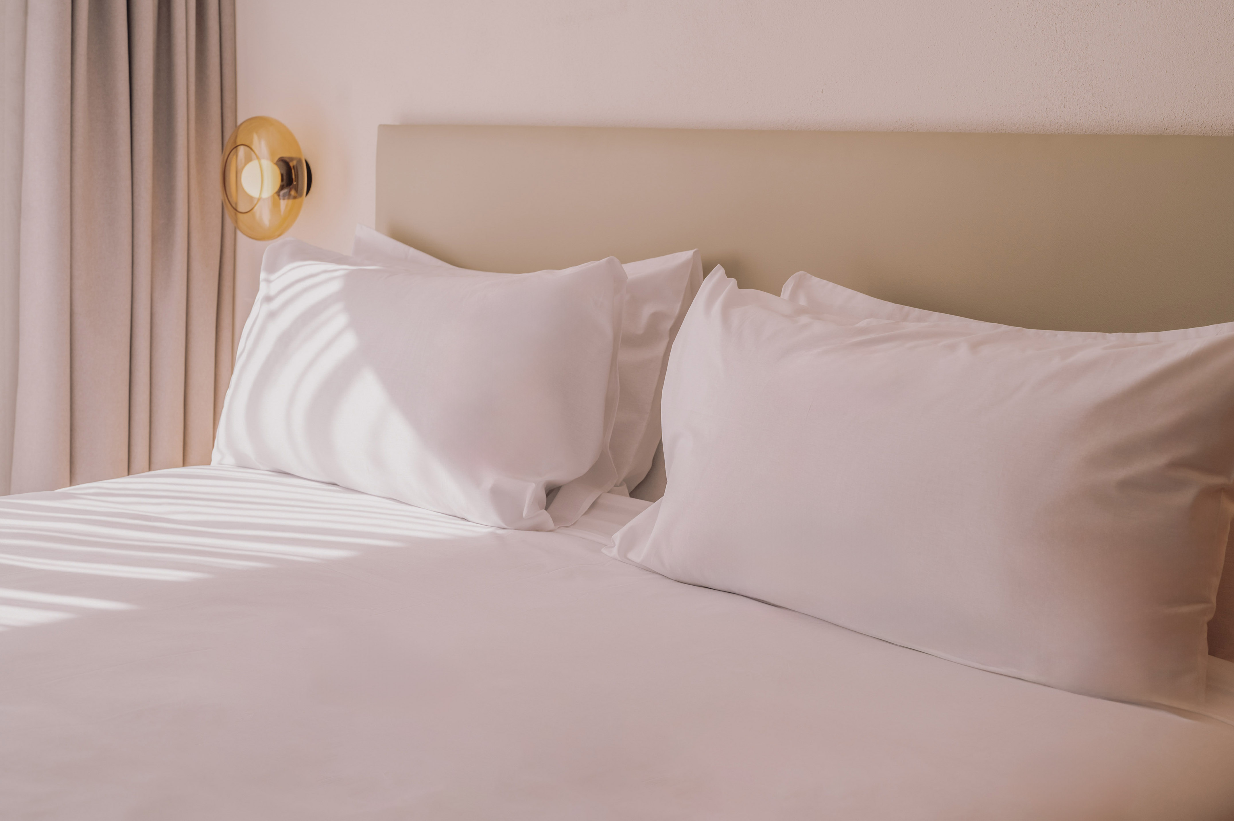 Hotel bed with white pillows, beige headboard, and amber wall lamp, sunlit.