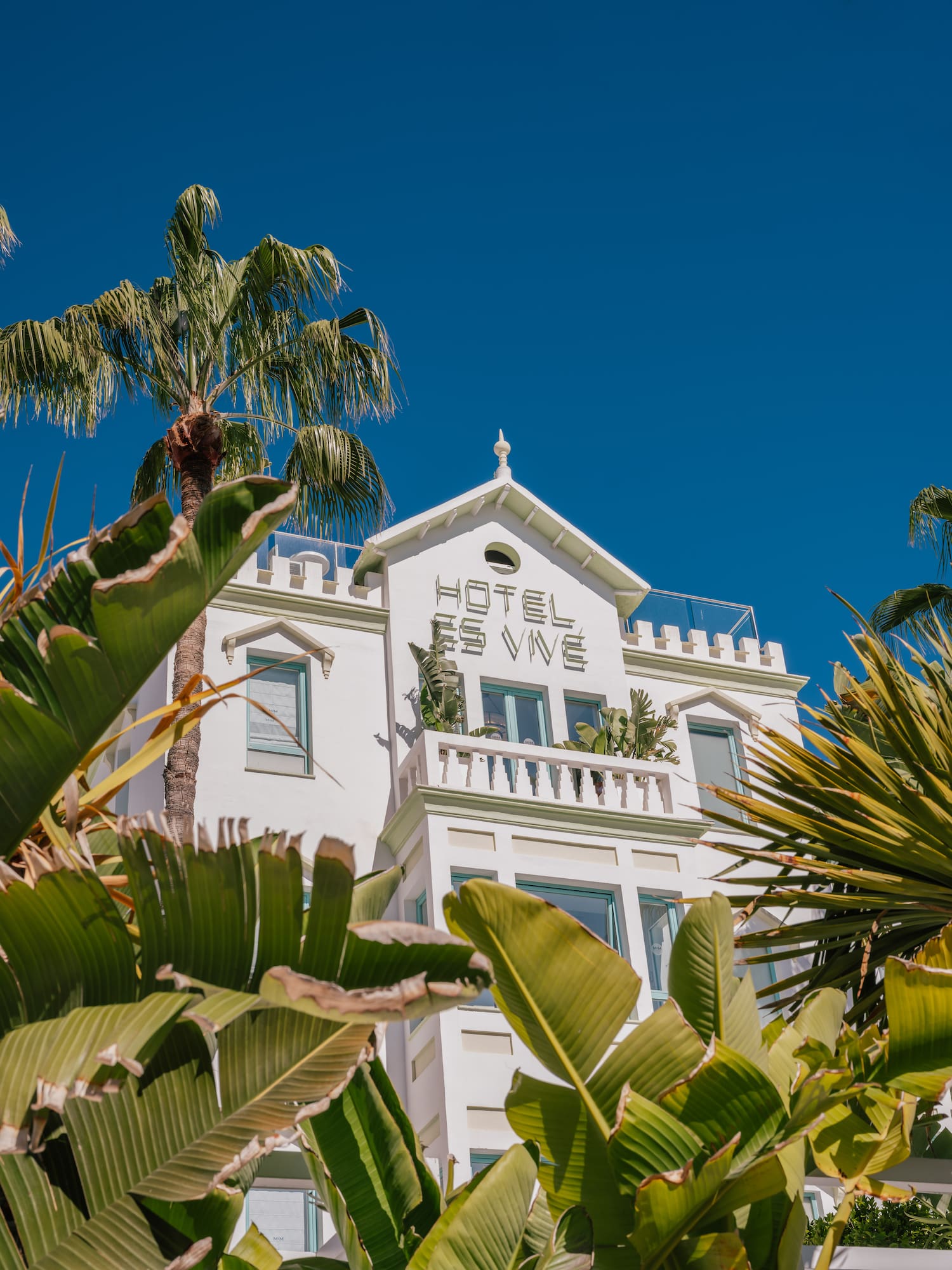 Hotel Es Vivé, white architecture among tropical palm trees and blue sky.