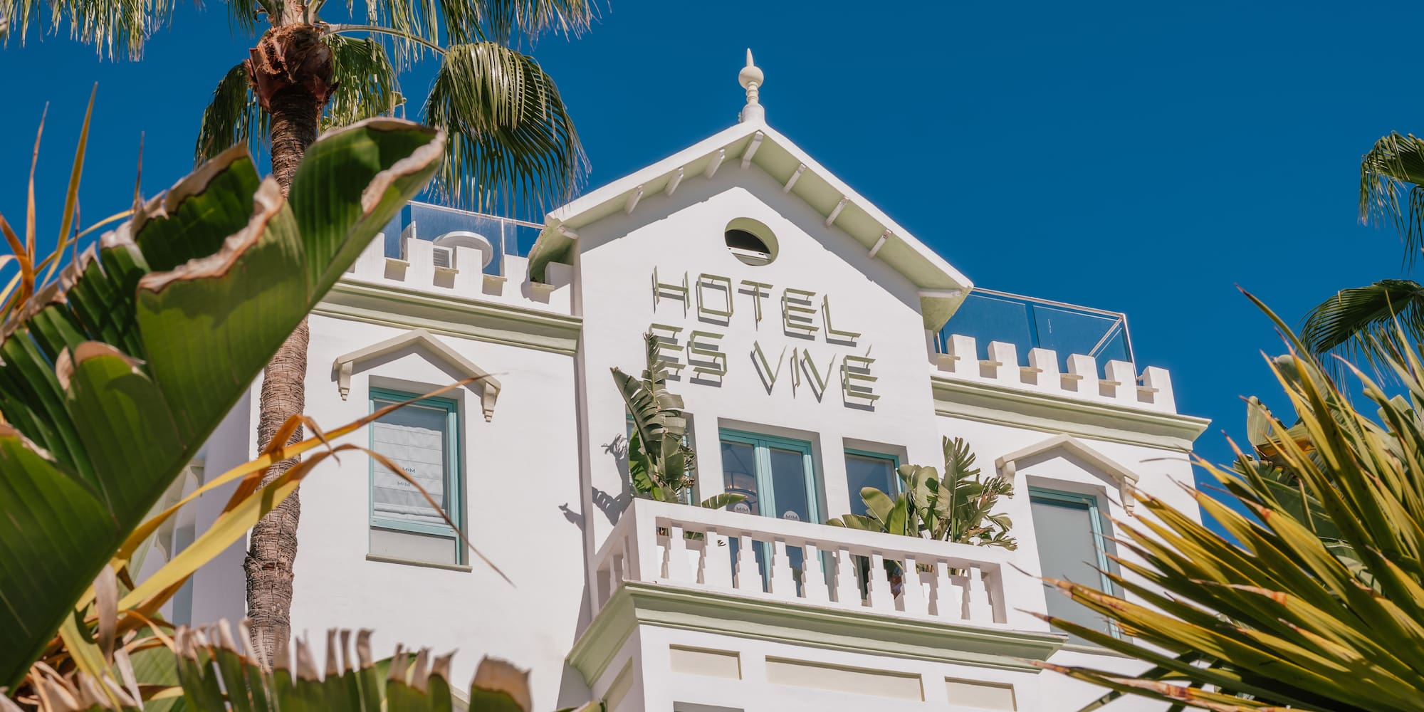 Hotel Es Vive Ibiza: white building, tropical palm trees, and clear blue sky.