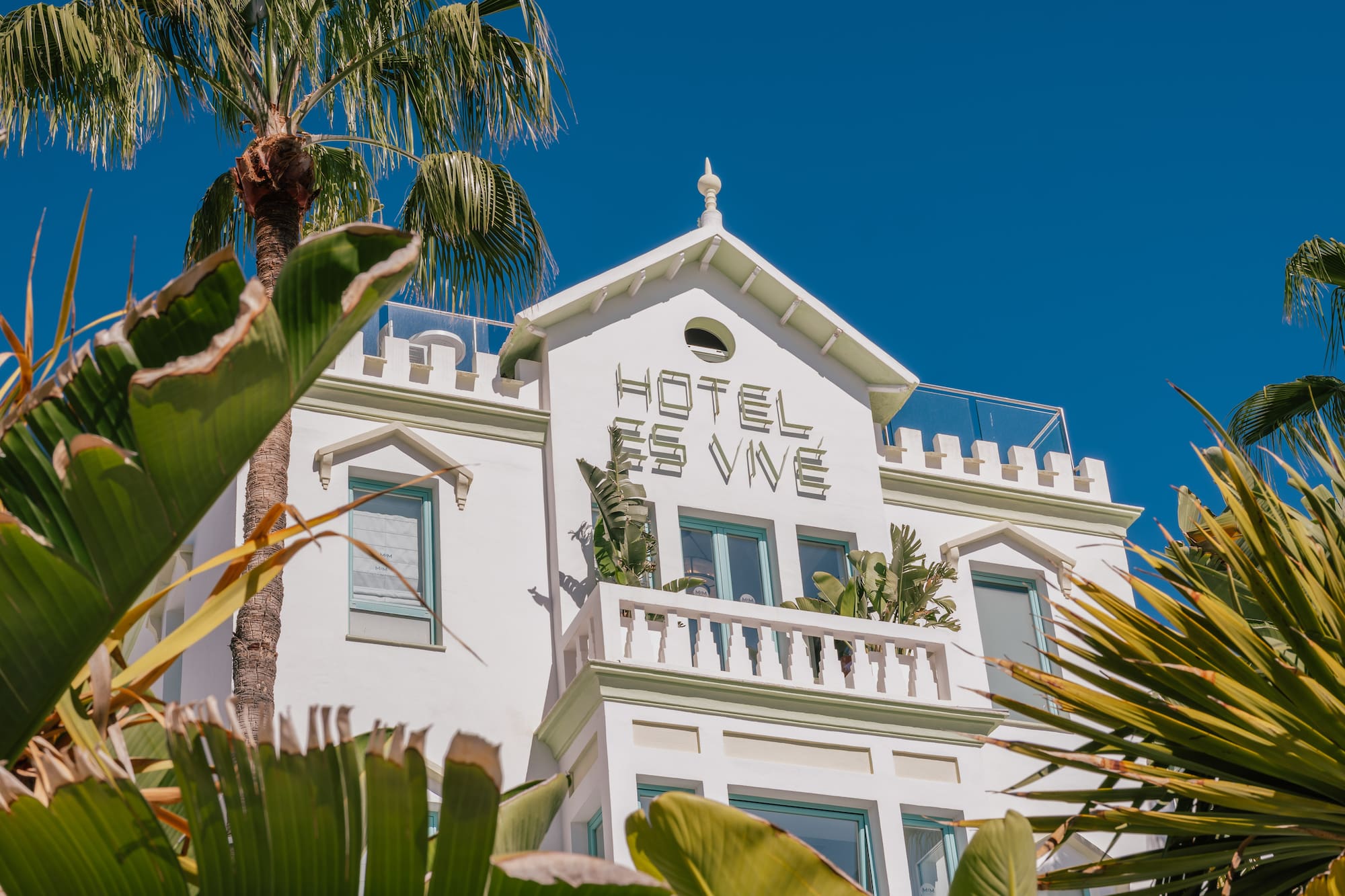 Hotel Es Vive, white building, green trim, palm trees, and tropical plants under blue sky.