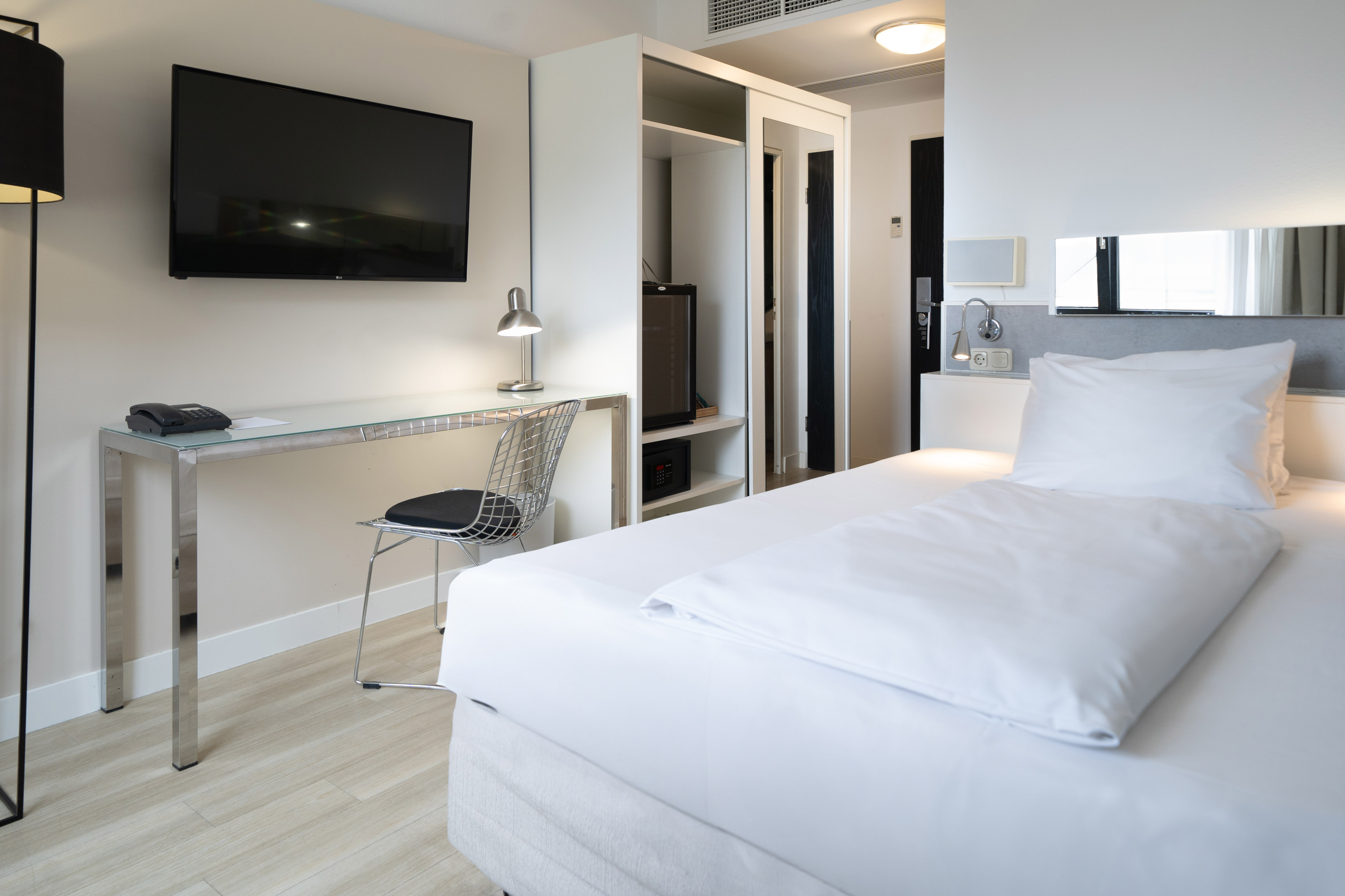 Modern, clean hotel room with white bed, desk, wall-mounted TV, and closet.