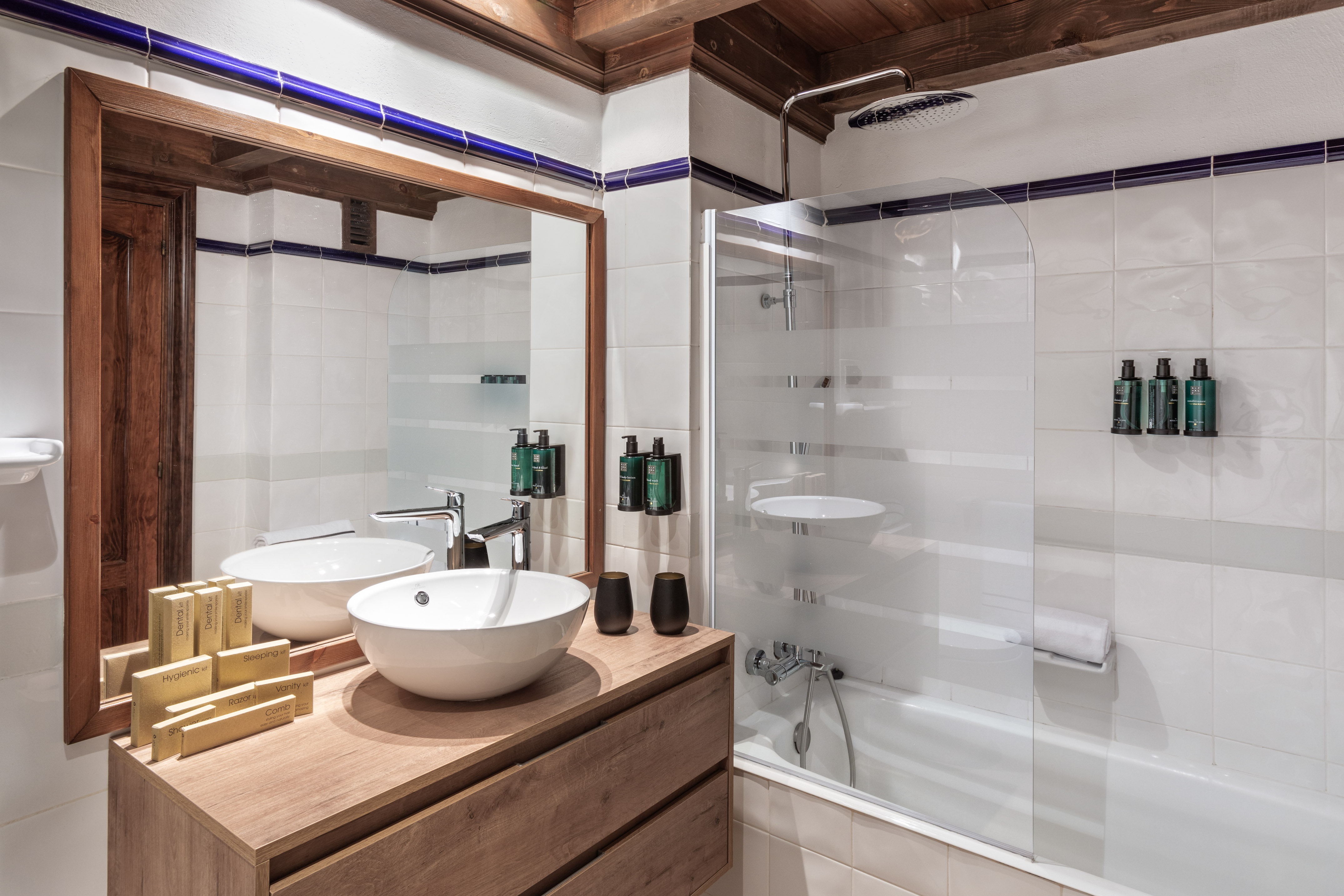 Hotel bathroom with wooden vanity, two vessel sinks, mirror, white tiles, and shower-tub.