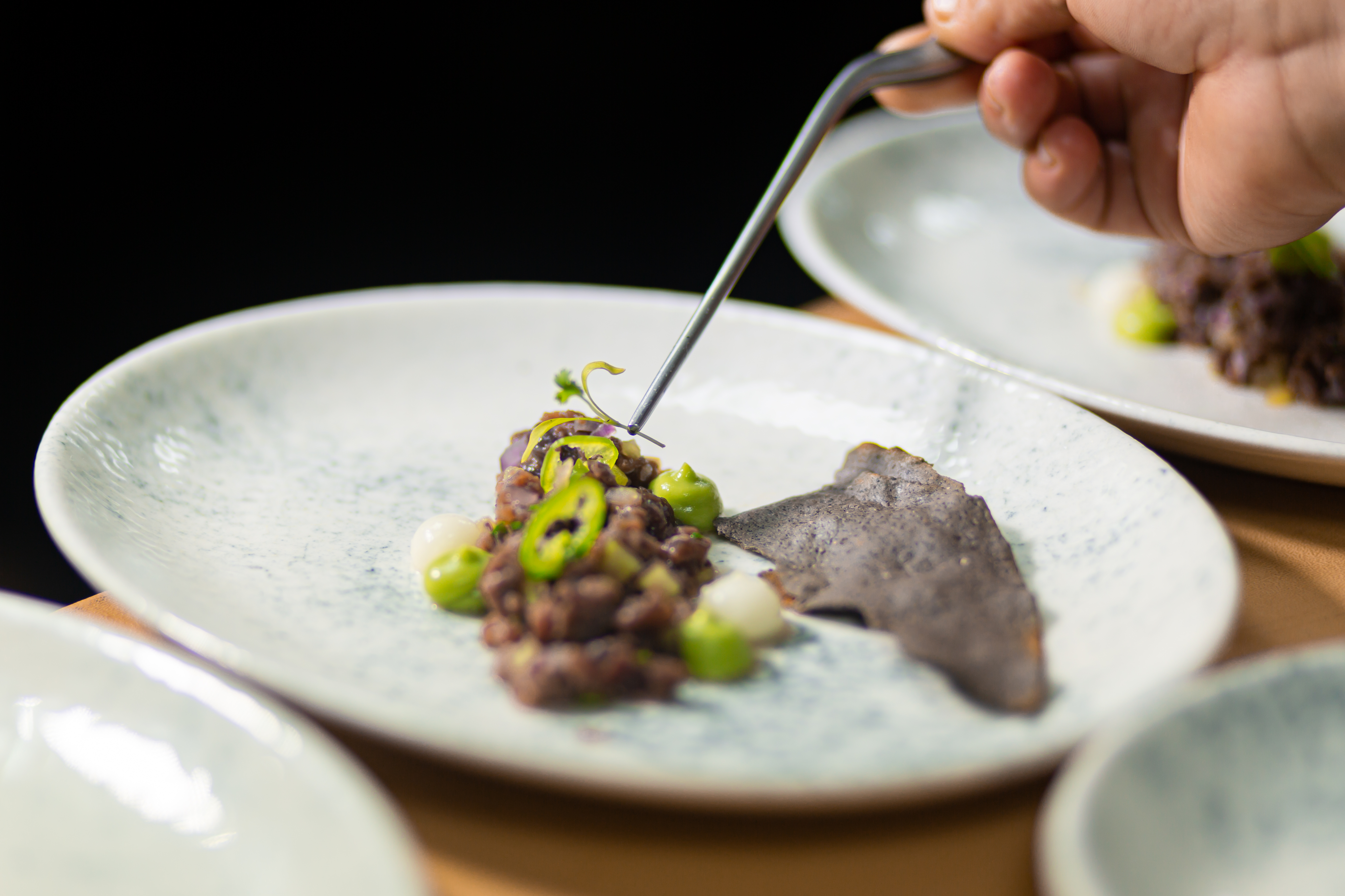 Chef plating fine dining dish with tweezers, adding garnish to food and crisps.