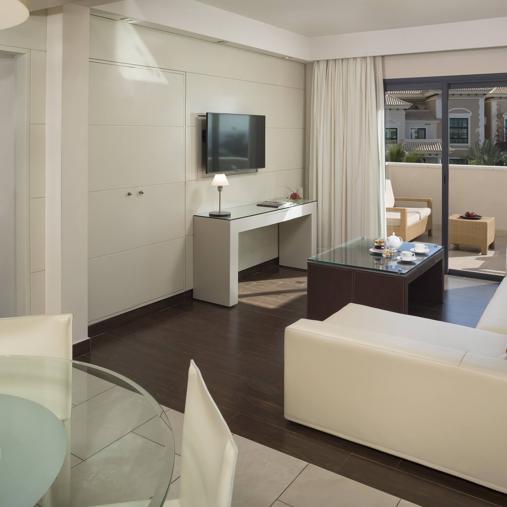 Modern hotel suite living room, cream sofa, TV, dining area, balcony.