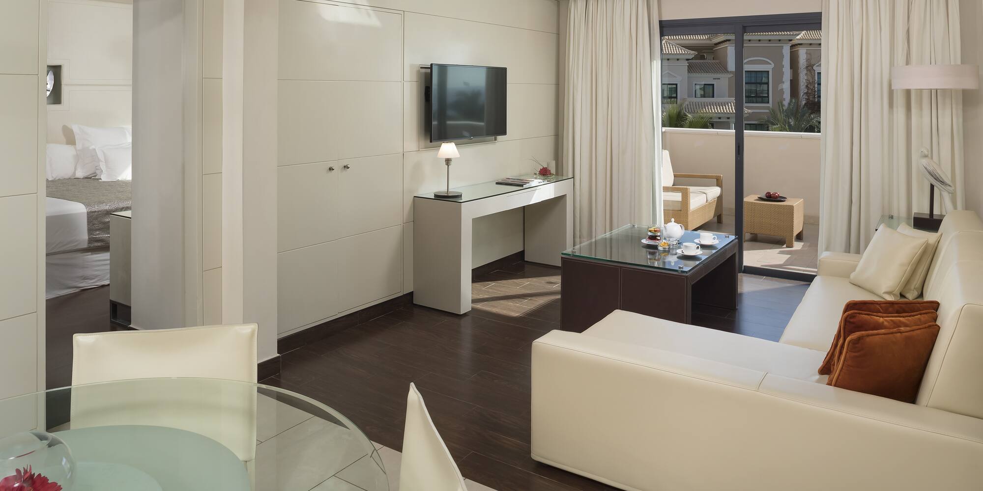 Modern hotel suite living room, cream sofa, TV, dining area, balcony.