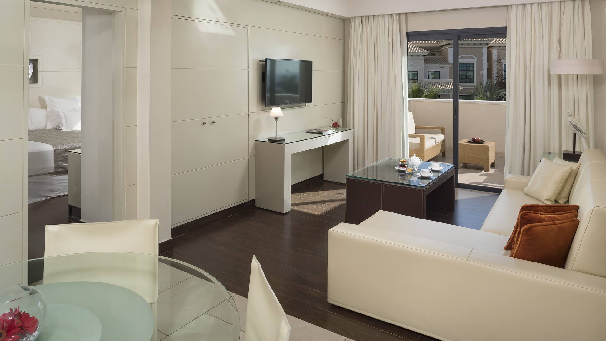 Modern hotel suite living room, cream sofa, TV, dining area, balcony.