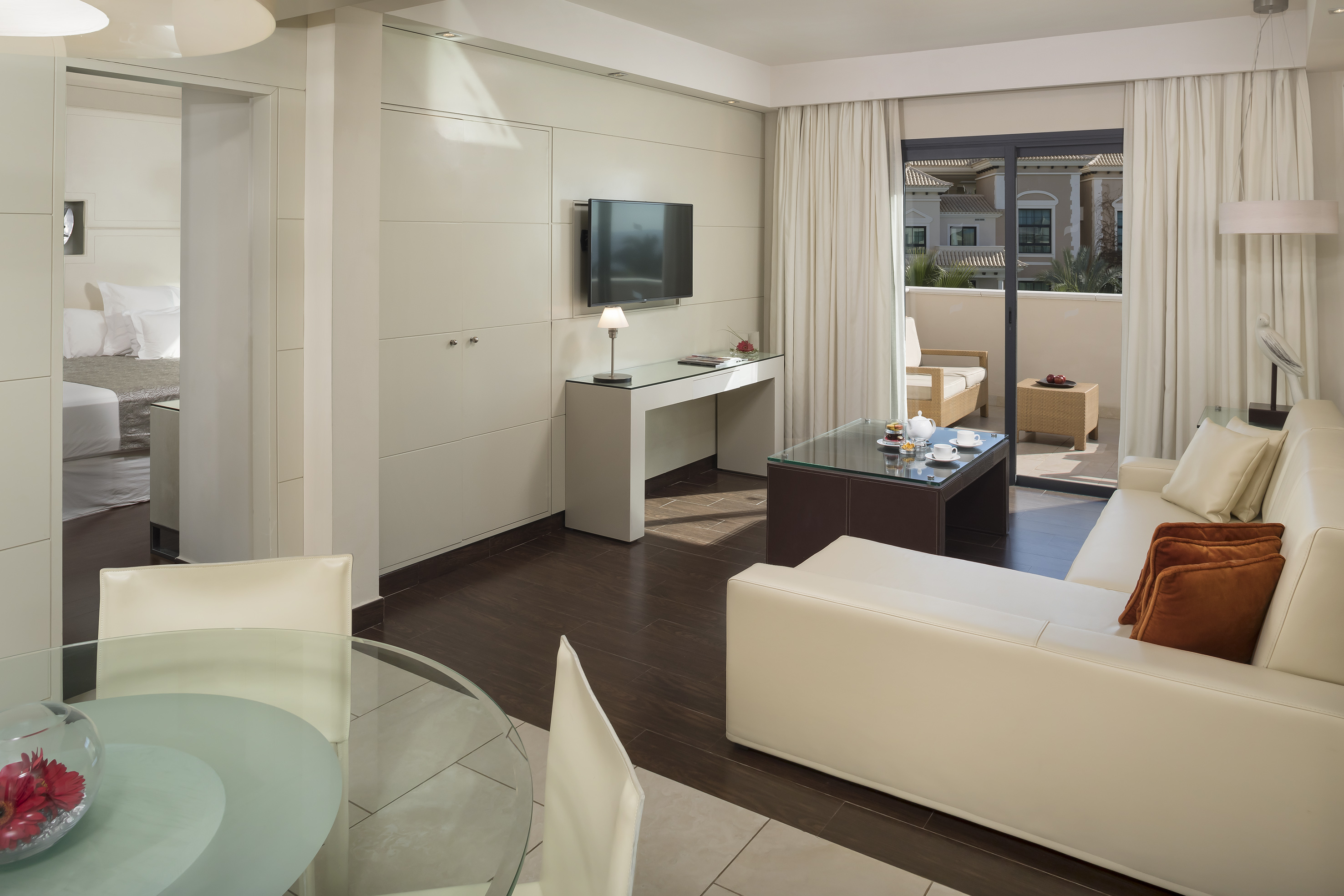 Modern hotel suite living room, cream sofa, TV, dining area, balcony.