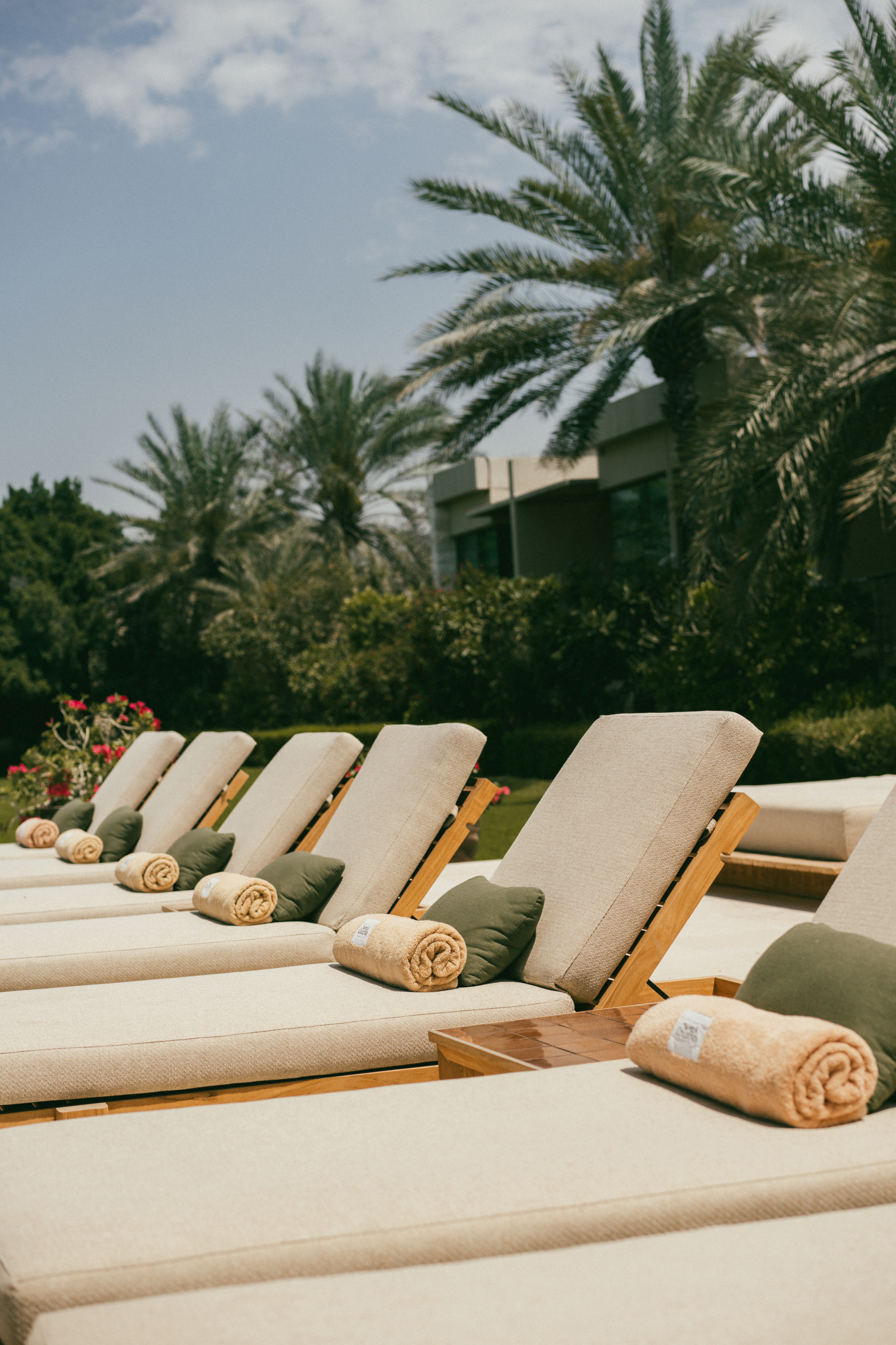 Resort sun loungers with green pillows, rolled towels, and palm trees.