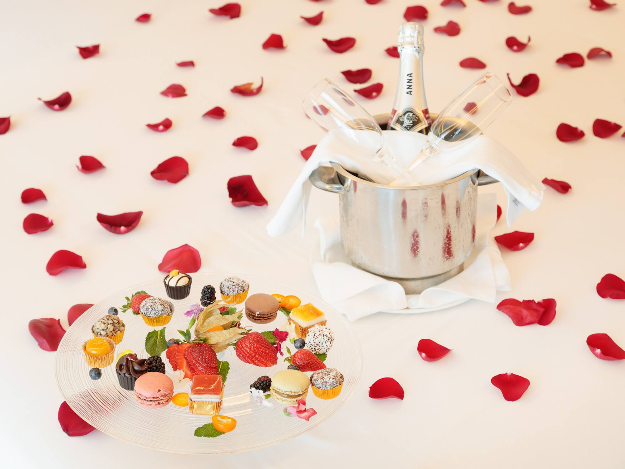 Champagne, desserts, and rose petals on white, for a romantic celebration.