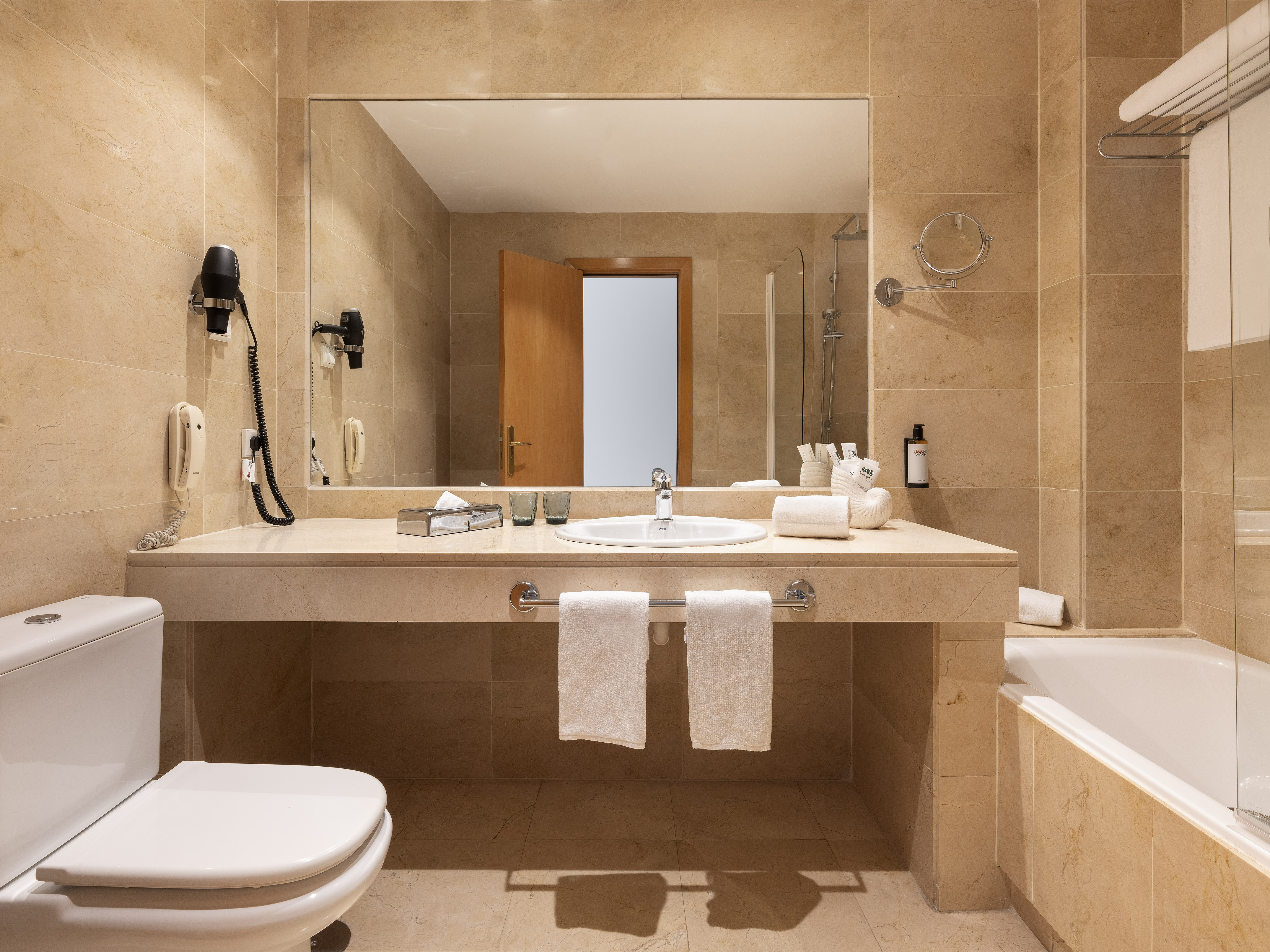 Tan marble hotel bathroom with vanity, sink, toilet, bathtub, and mirror.