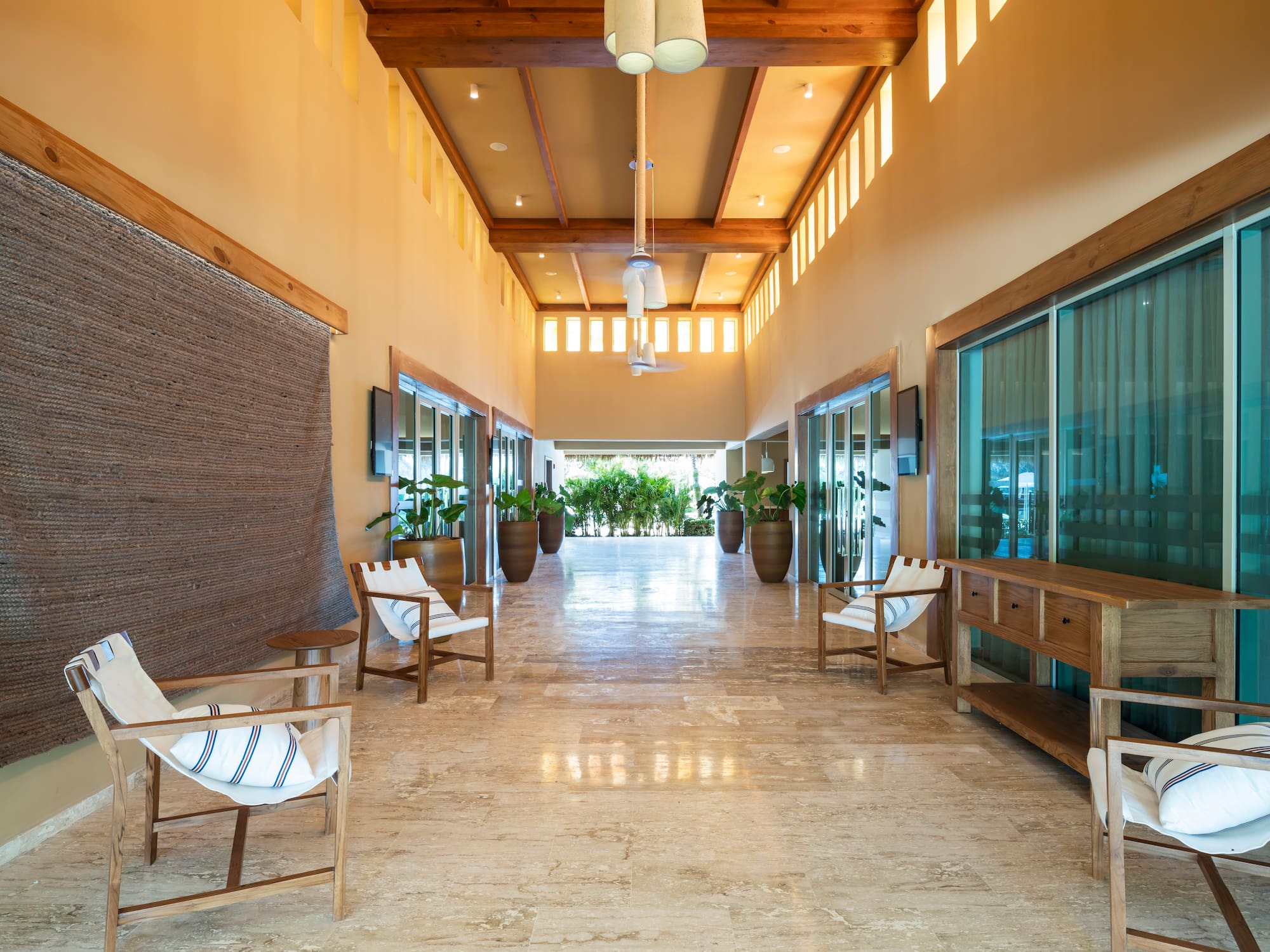 Hotel hallway with wood beams, tiled floor, large plants, and chairs.