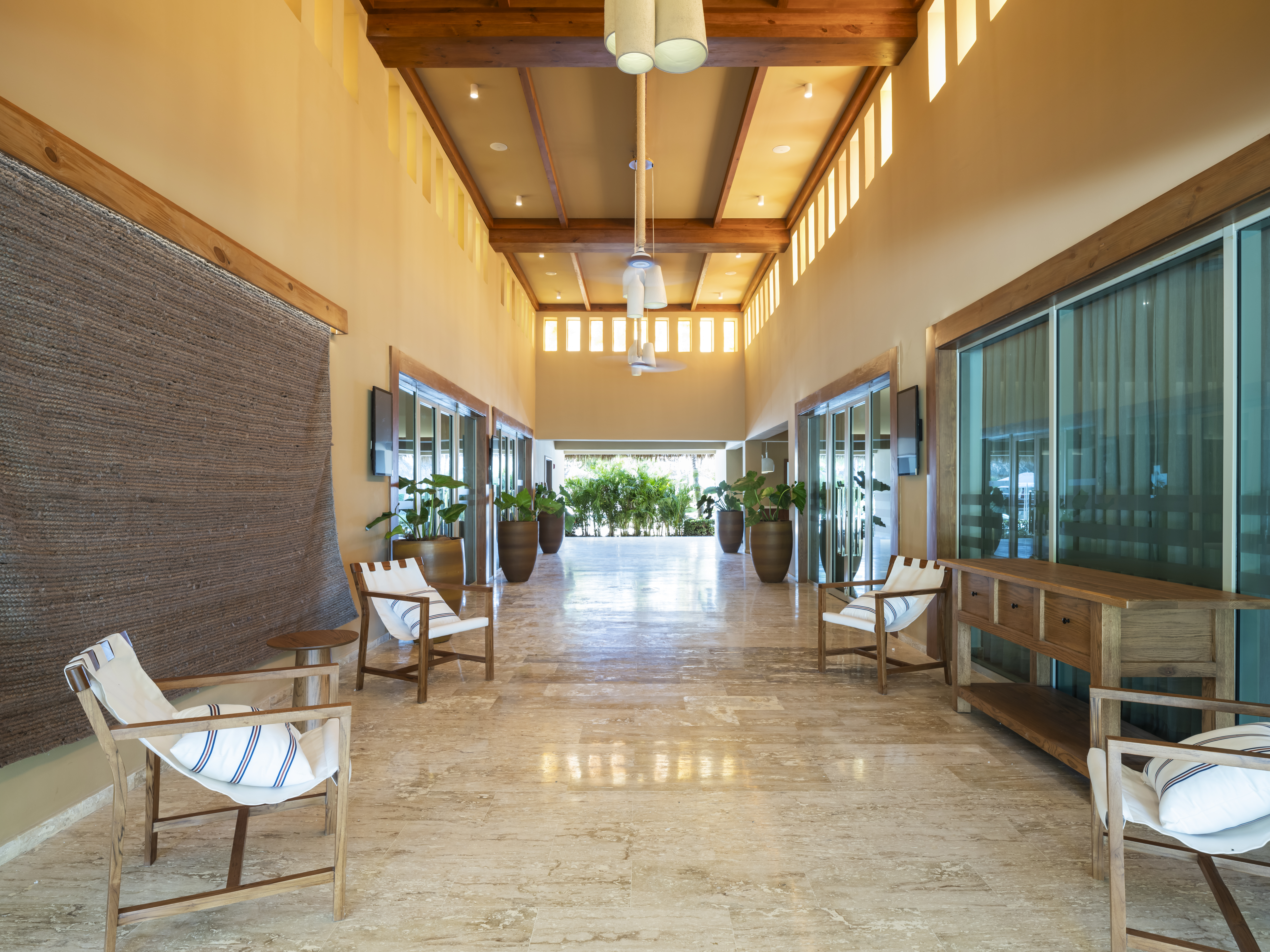 Hotel hallway with wood beams, tiled floor, large plants, and chairs.