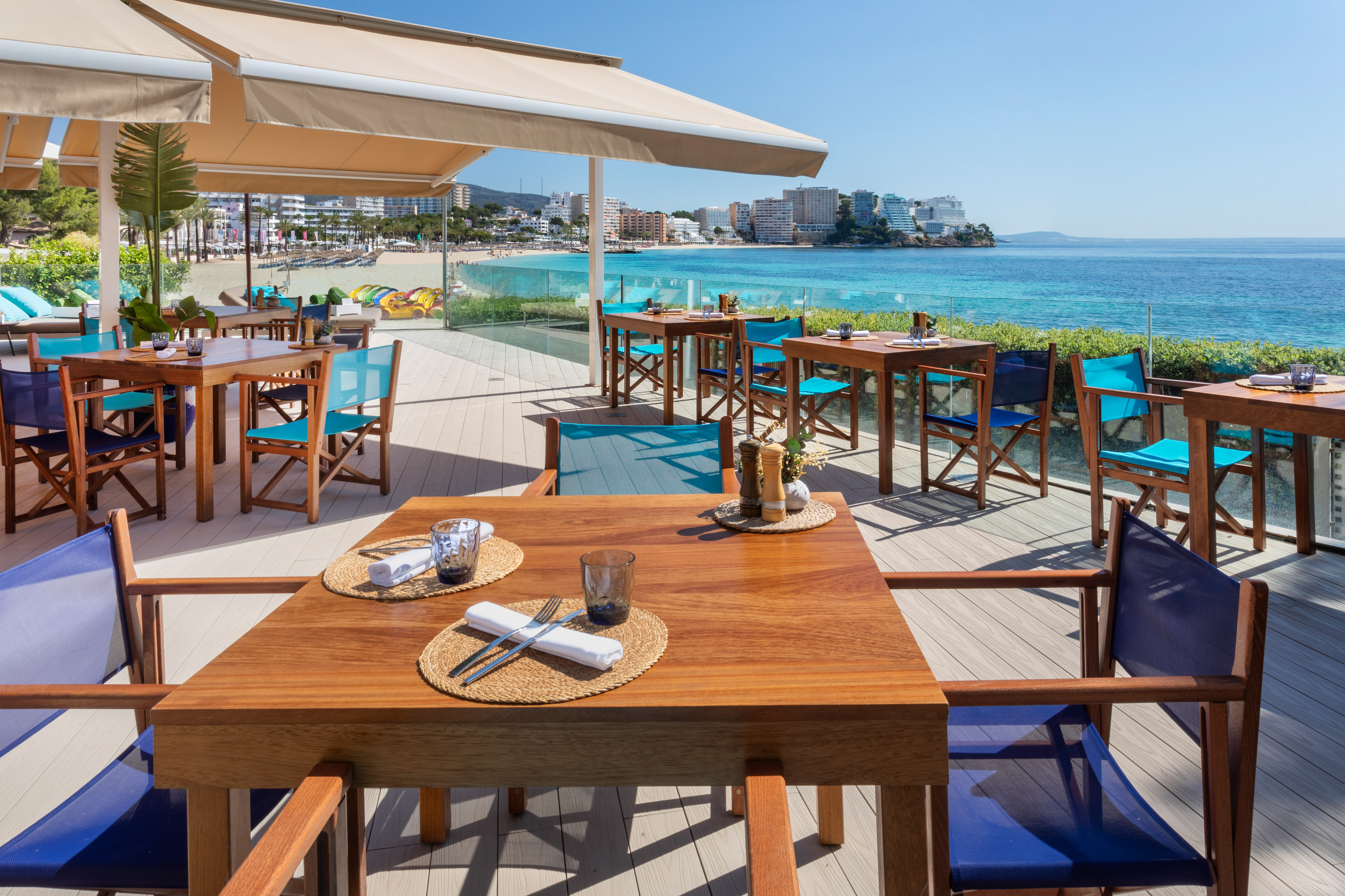 Beachfront restaurant terrace, wooden tables, blue chairs, turquoise sea.