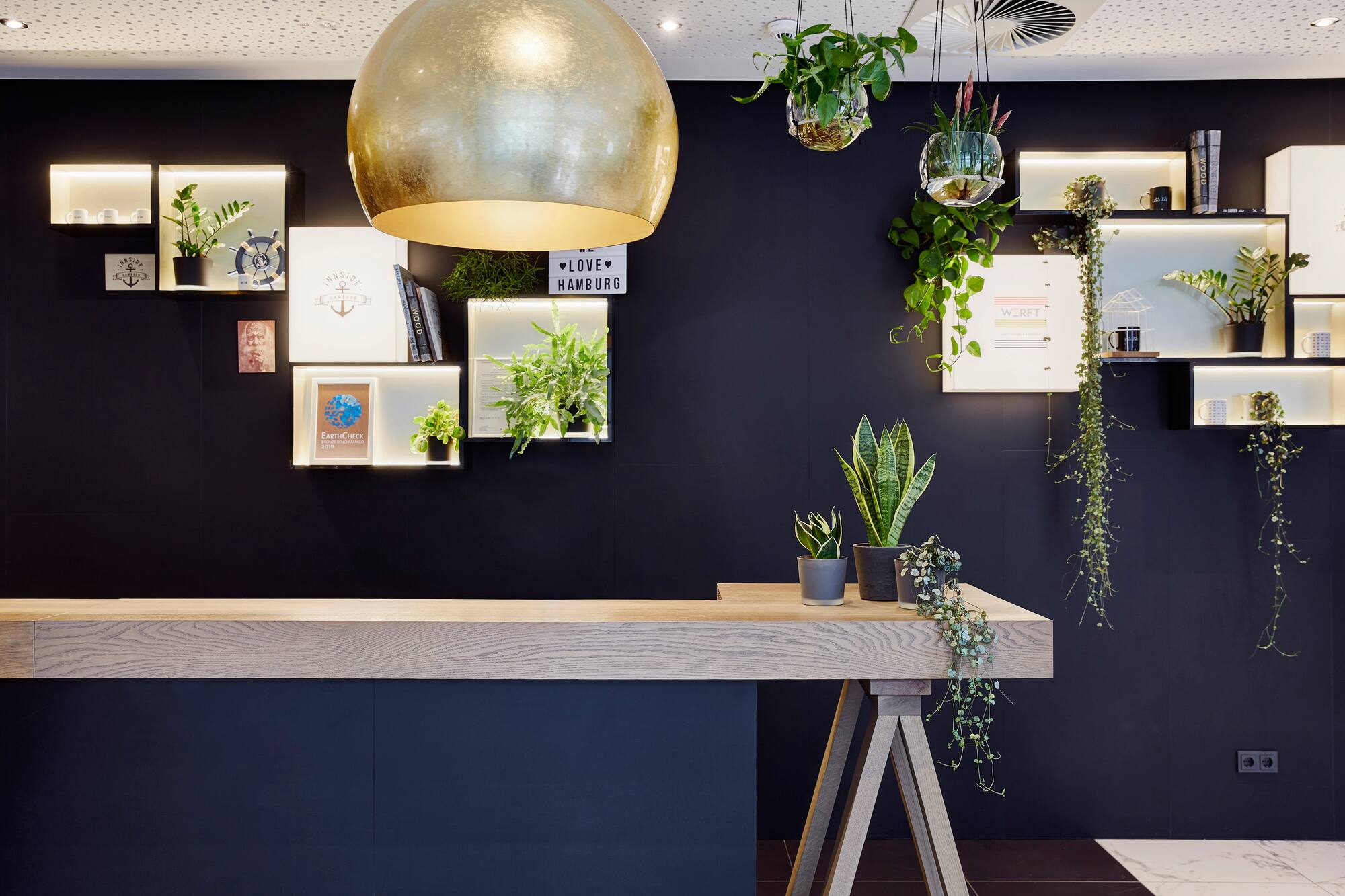 Modern hotel reception desk with dark wall, green plants, and golden light.