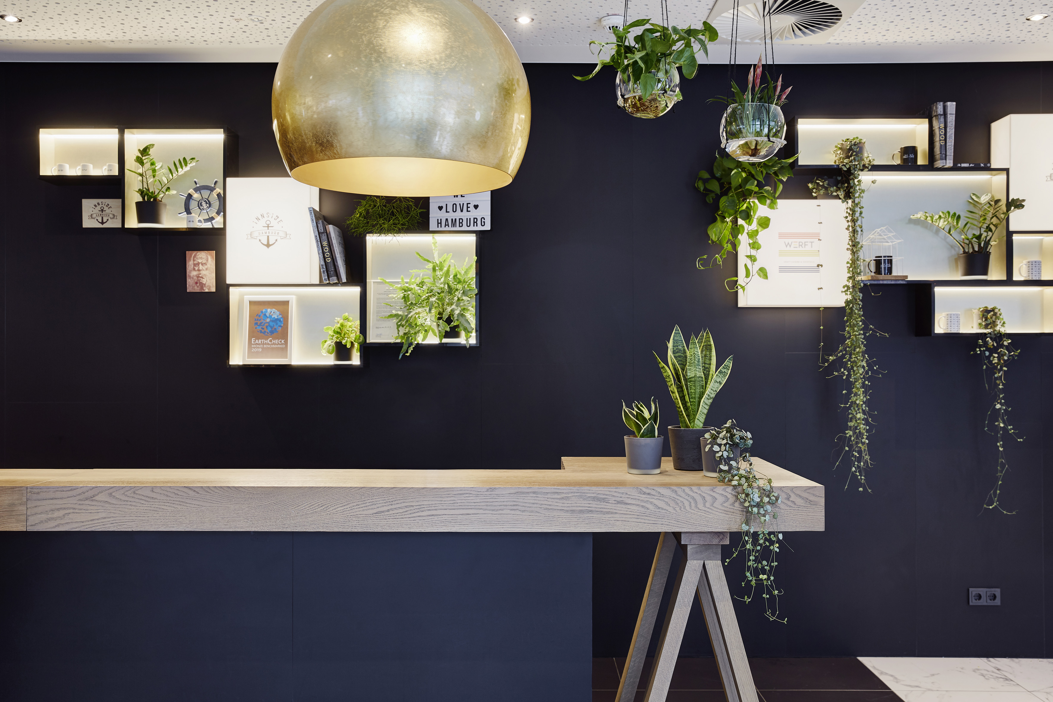 Modern hotel reception desk with dark wall, green plants, and golden light.