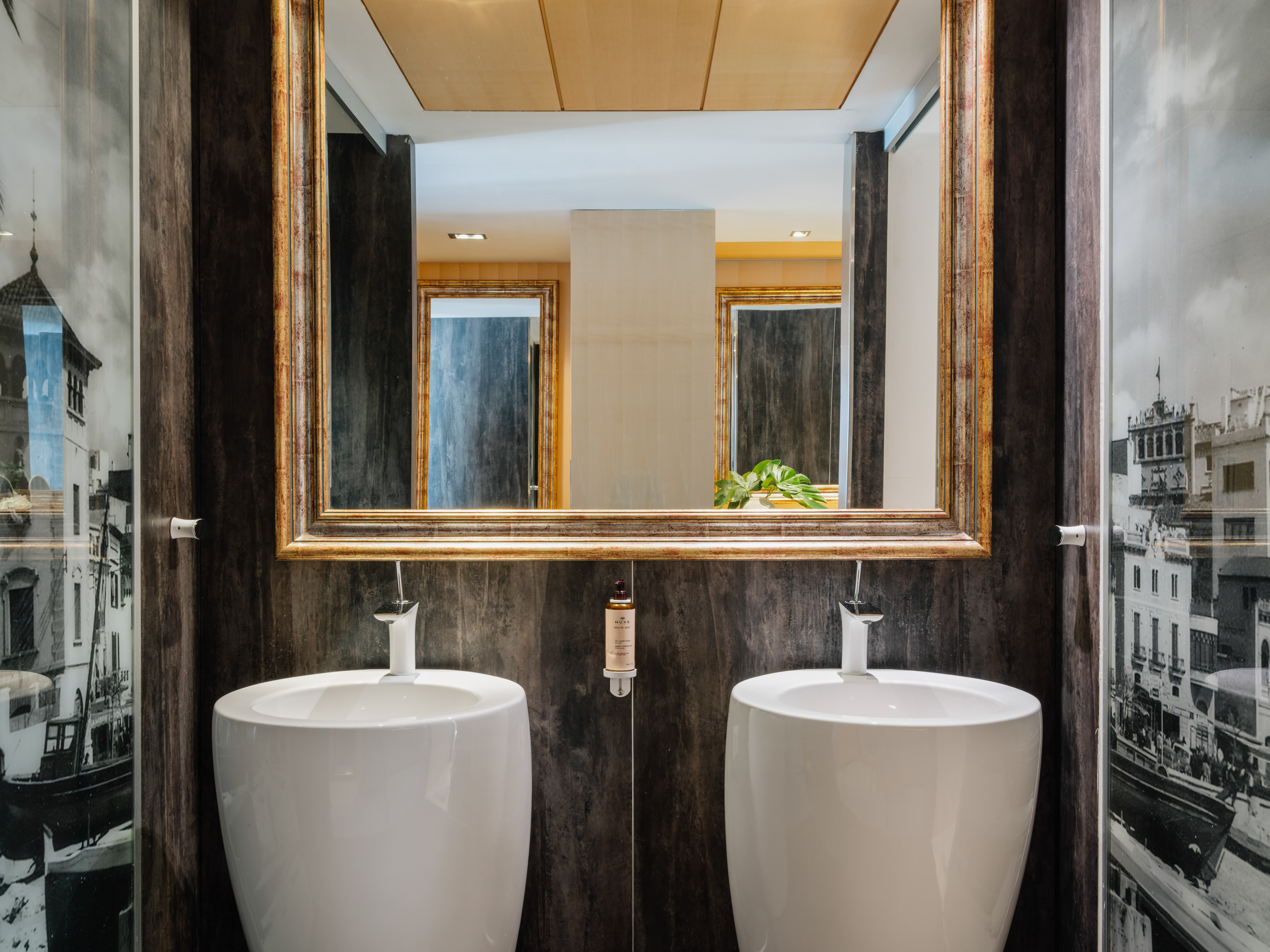 Modern bathroom with double pedestal sinks, gold mirror, dark walls.
