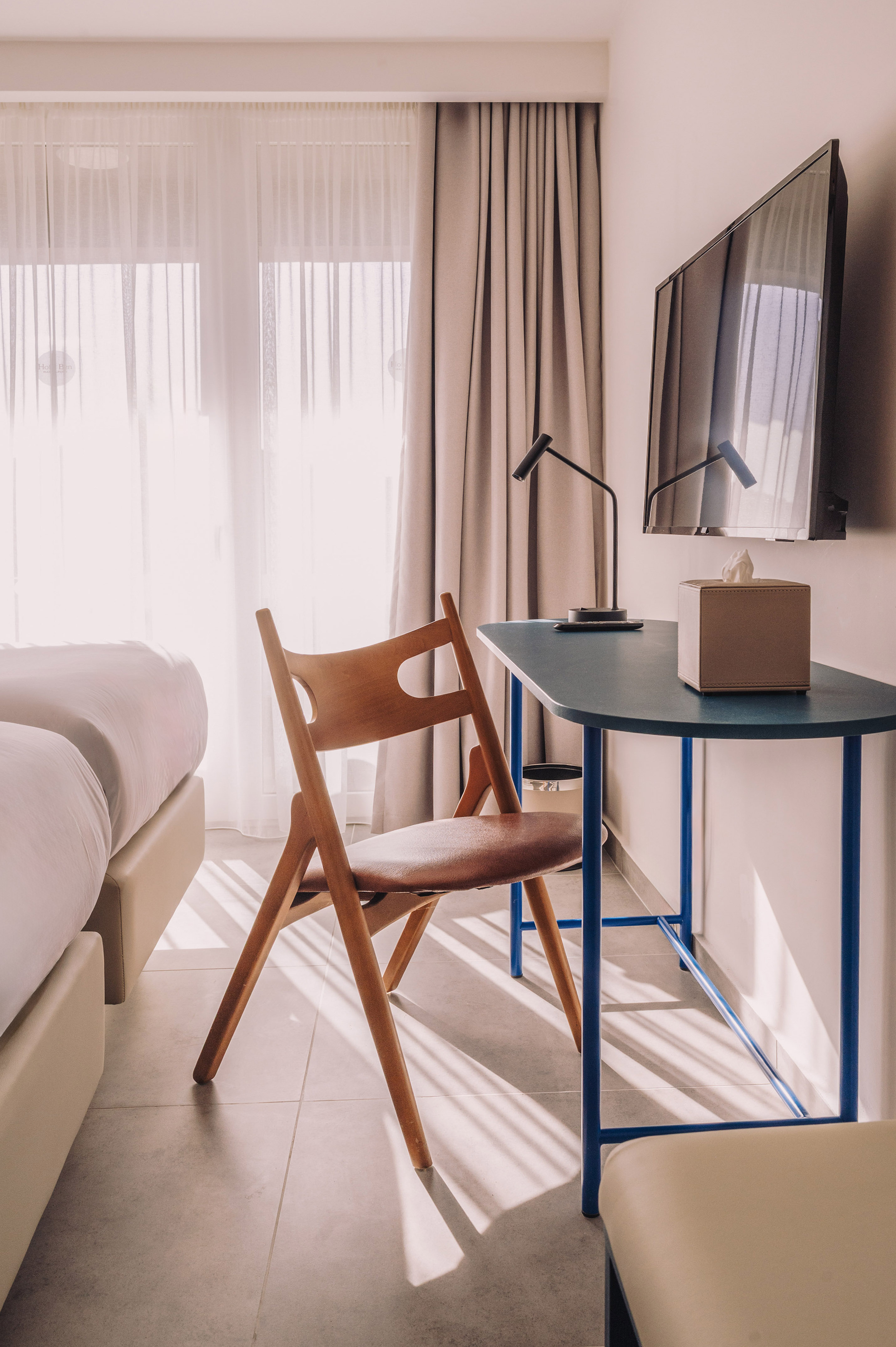 Modern hotel bedroom with a blue desk, wooden chair, wall TV, and sunny window.