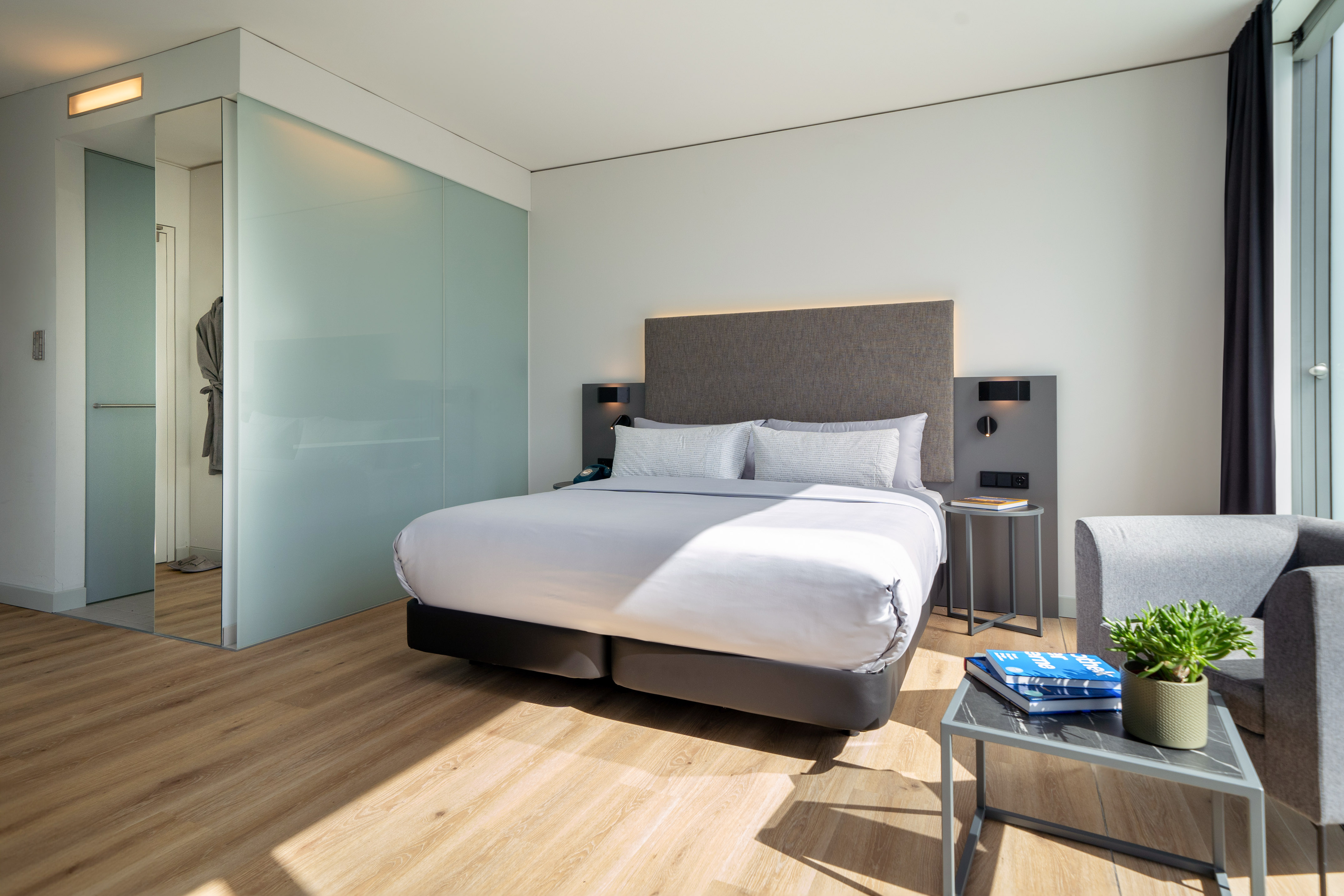 Contemporary hotel bedroom with king bed, wood floor, and glass partition.