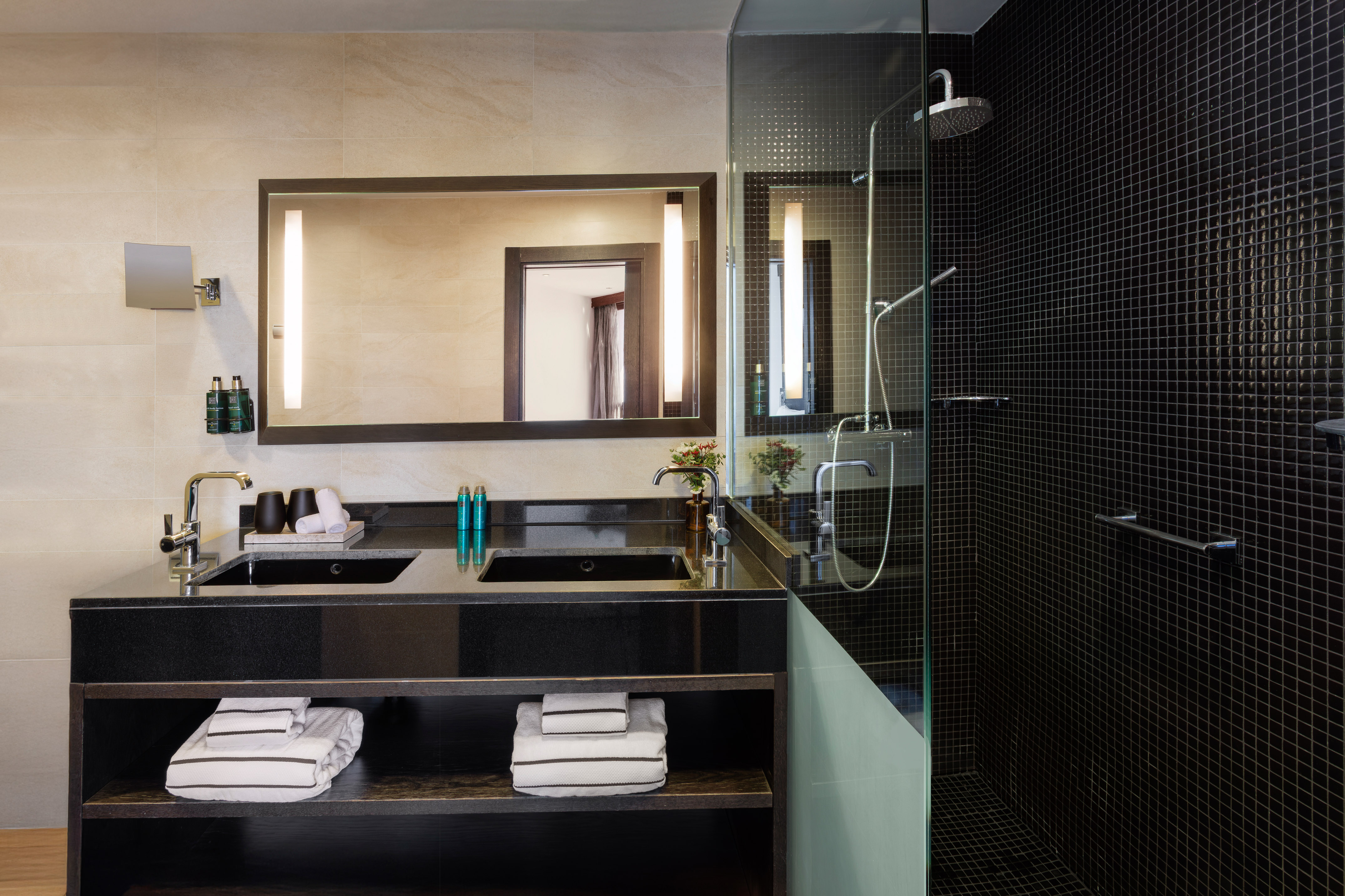 Modern hotel bathroom with black double vanity, lighted mirror, glass shower.