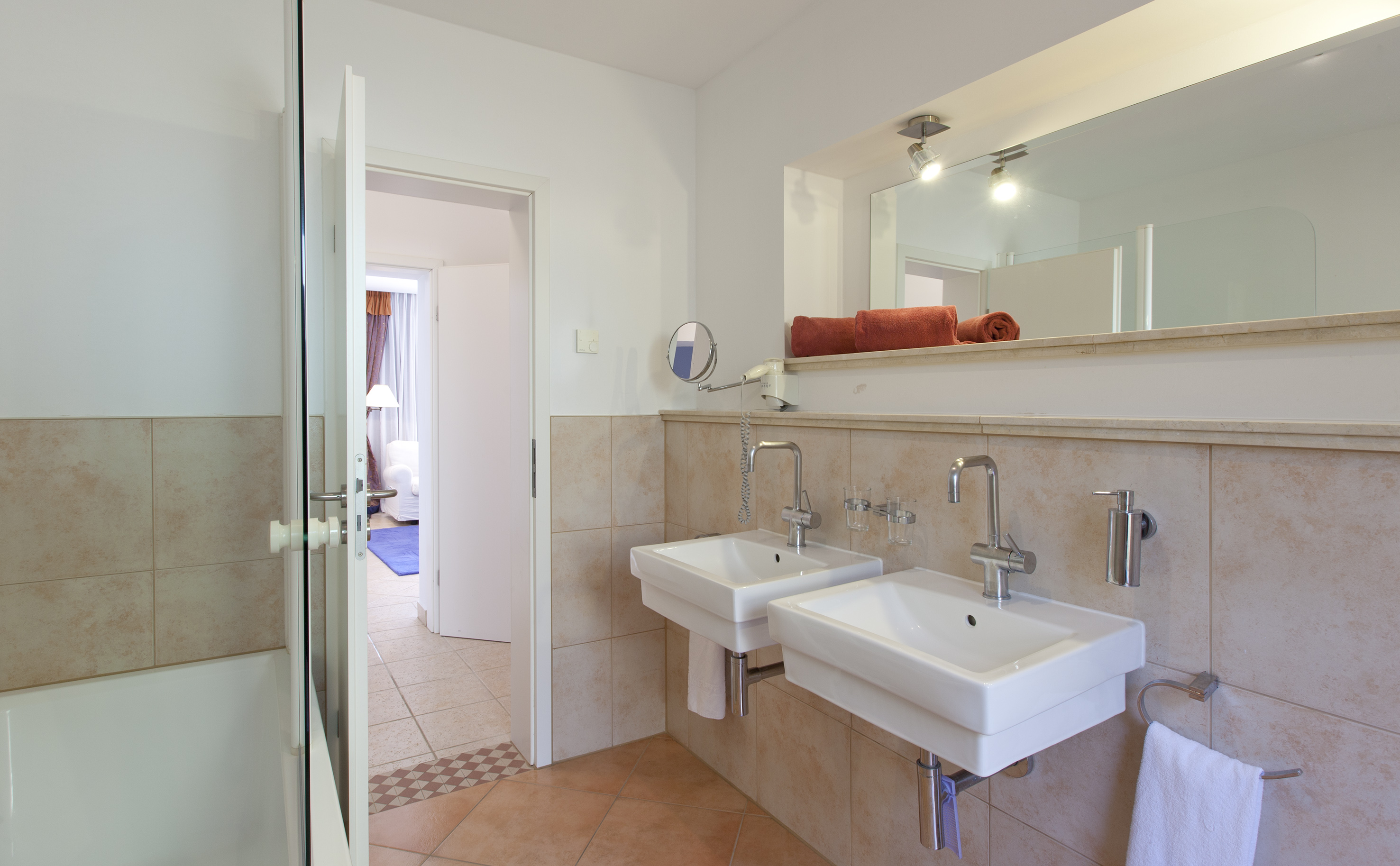 Hotel bathroom featuring dual white sinks, beige tiles, and large mirror.