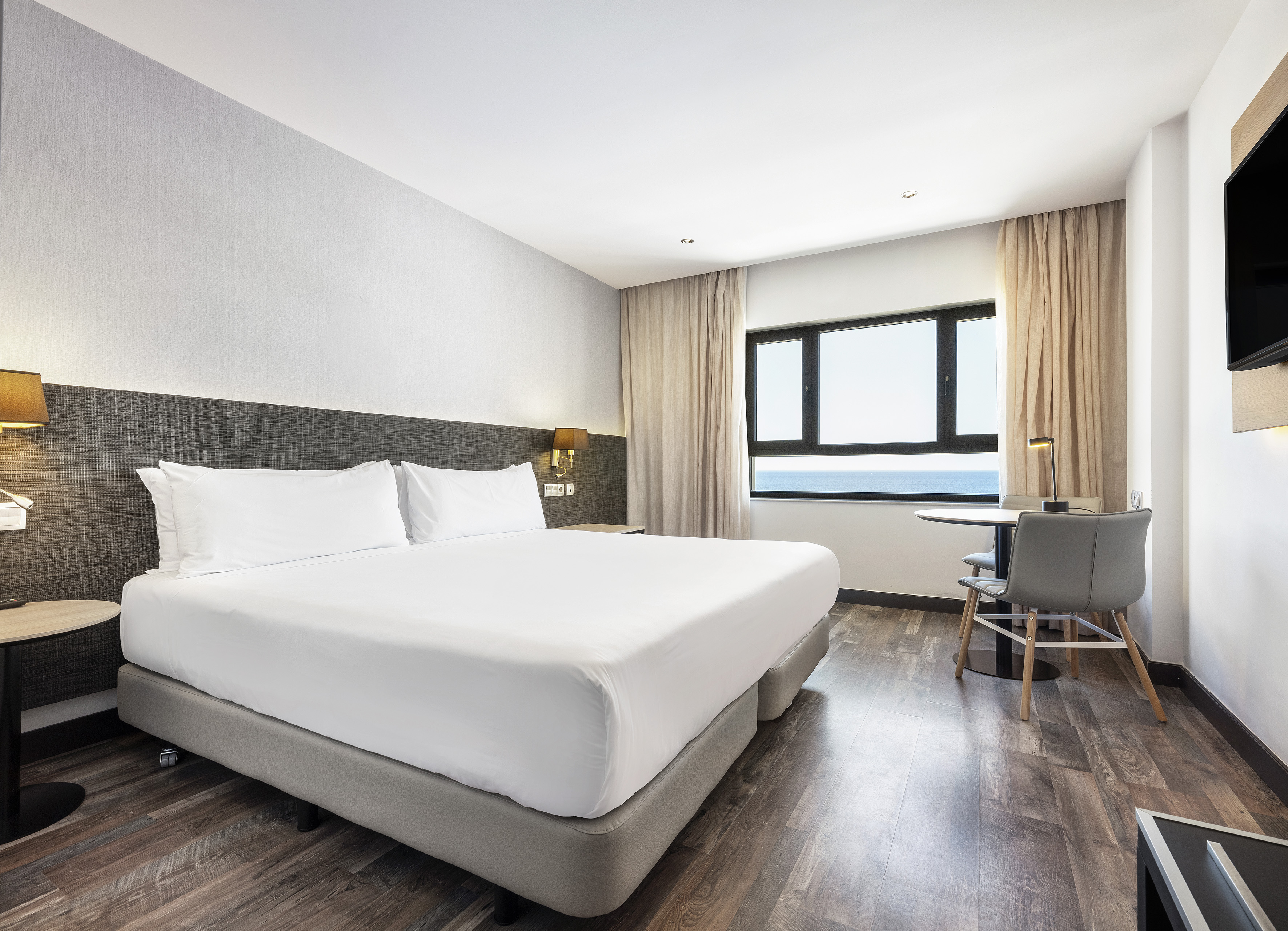 Modern hotel room with large bed, ocean view window, wooden floor, and work table.