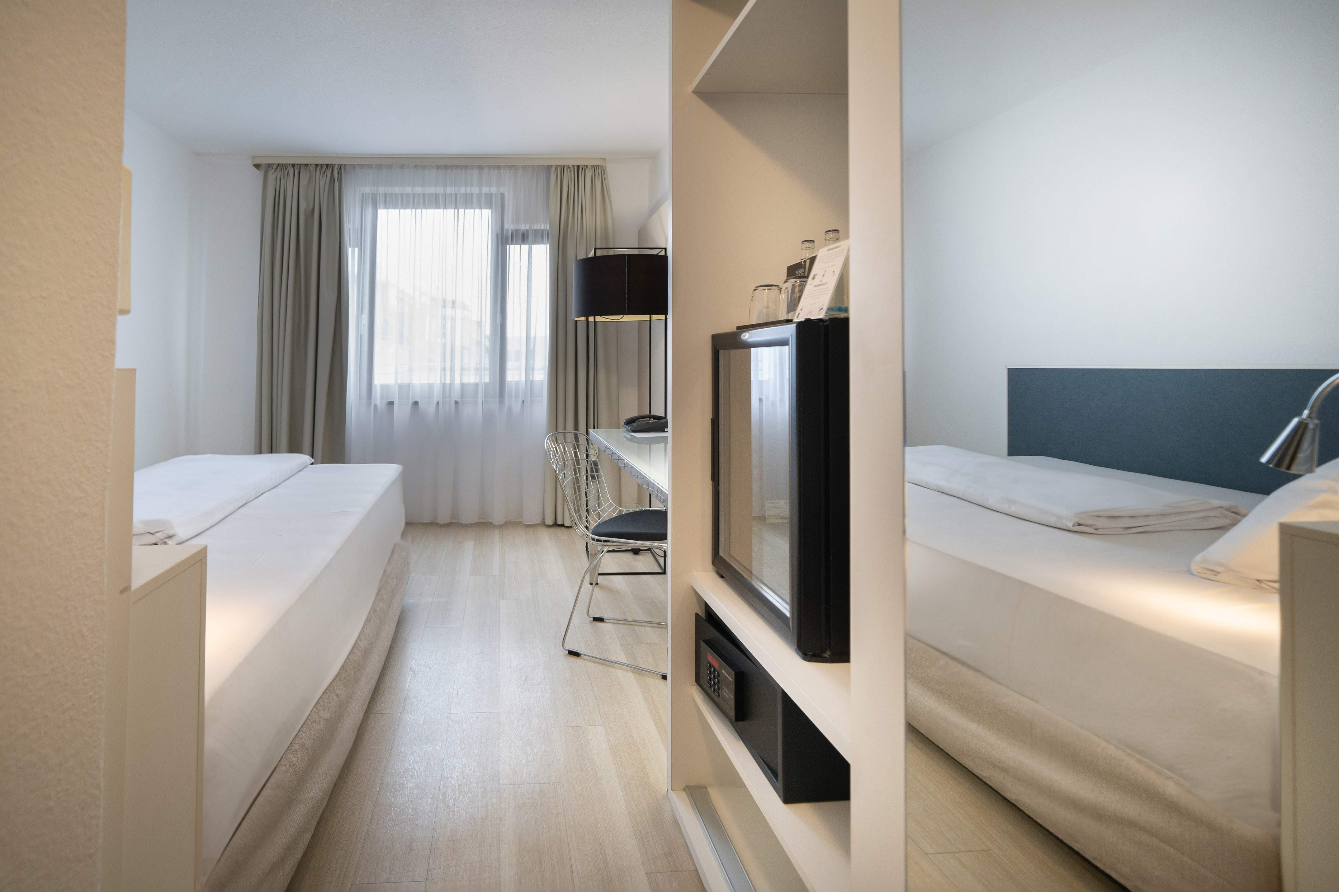 Hotel room with separate beds, desk, mini-fridge, and light wood floor.