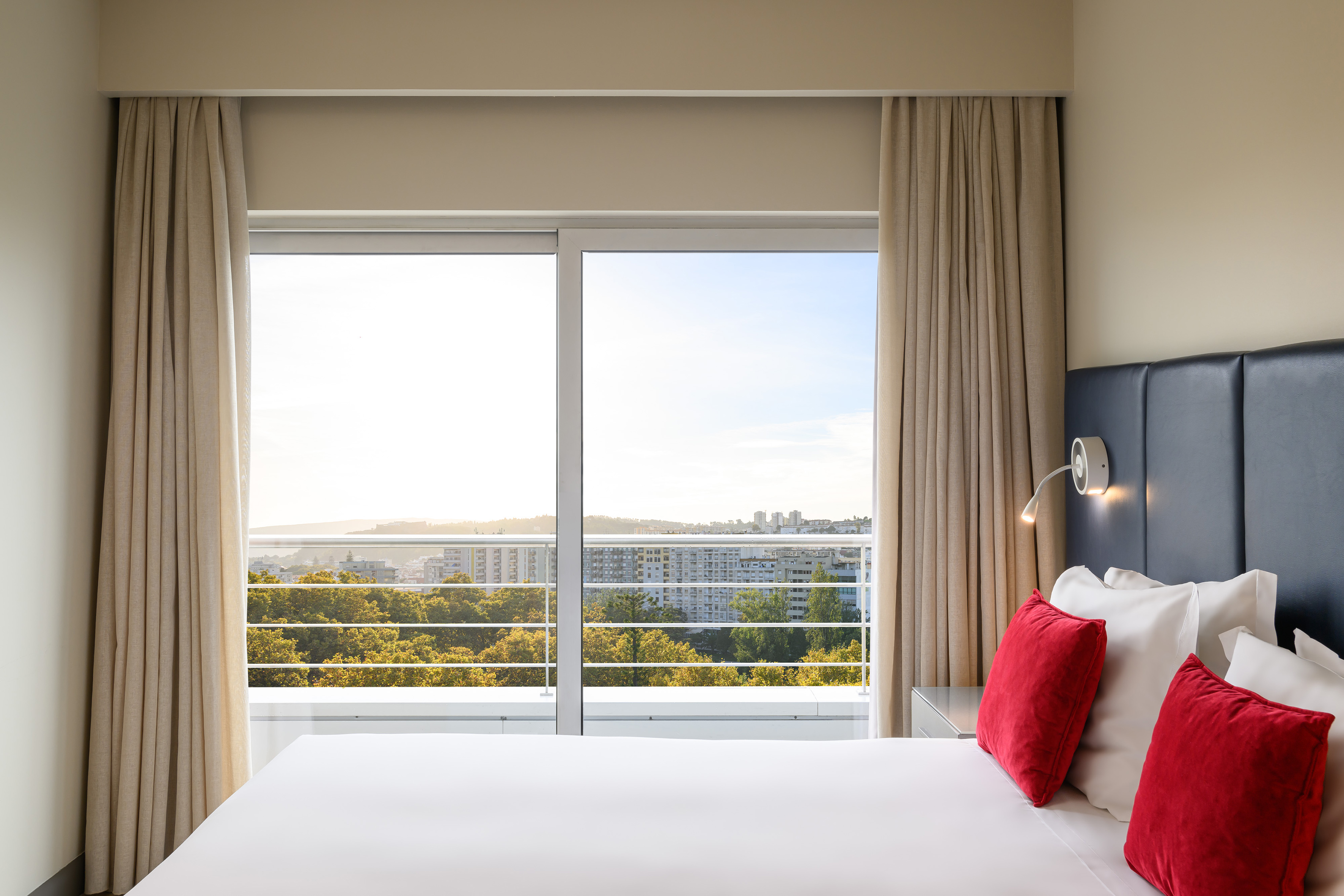 Hotel bedroom with white bed, red pillows, and city skyline view from window.