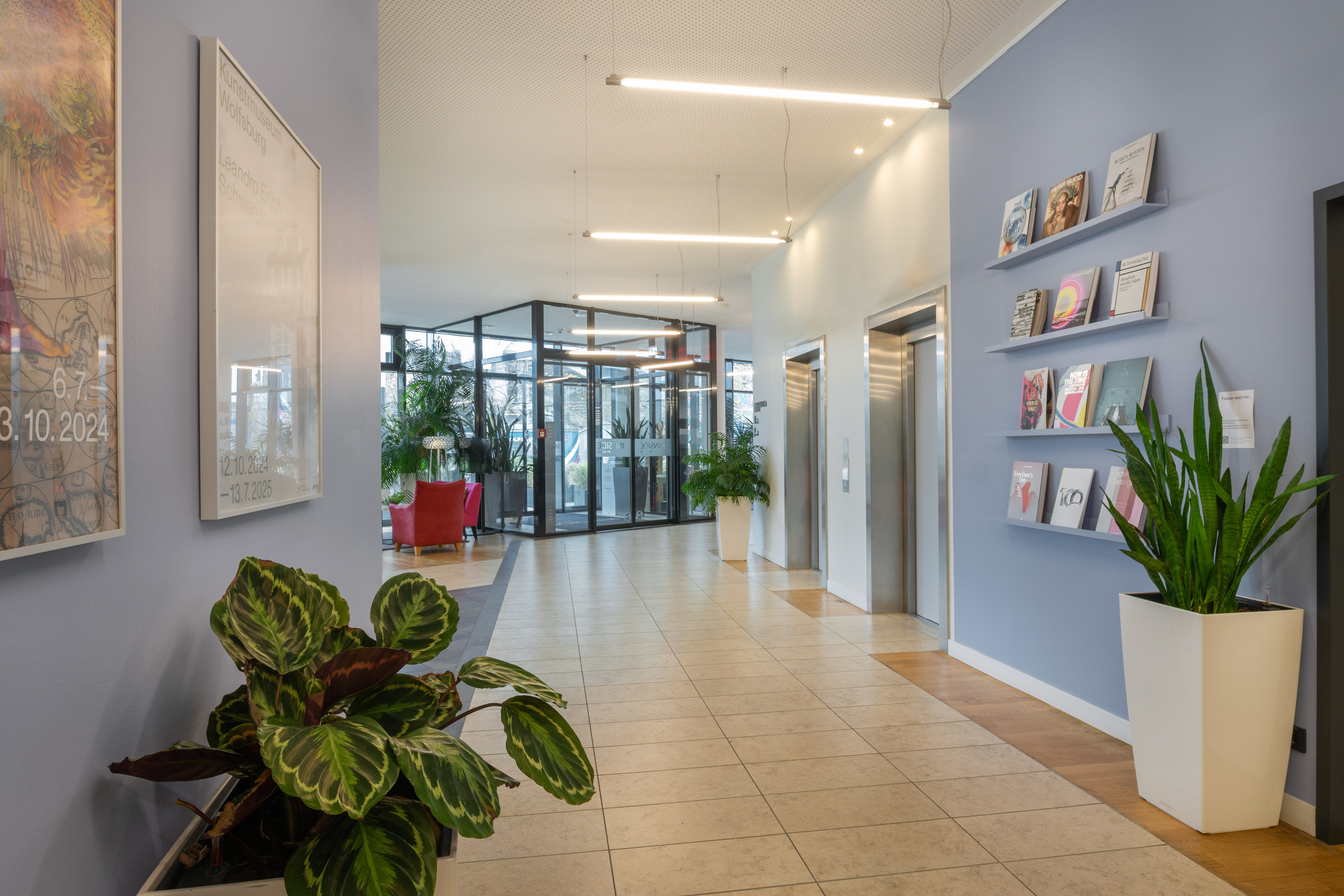 Bright modern office lobby with elevators, potted plants, and magazine displays.