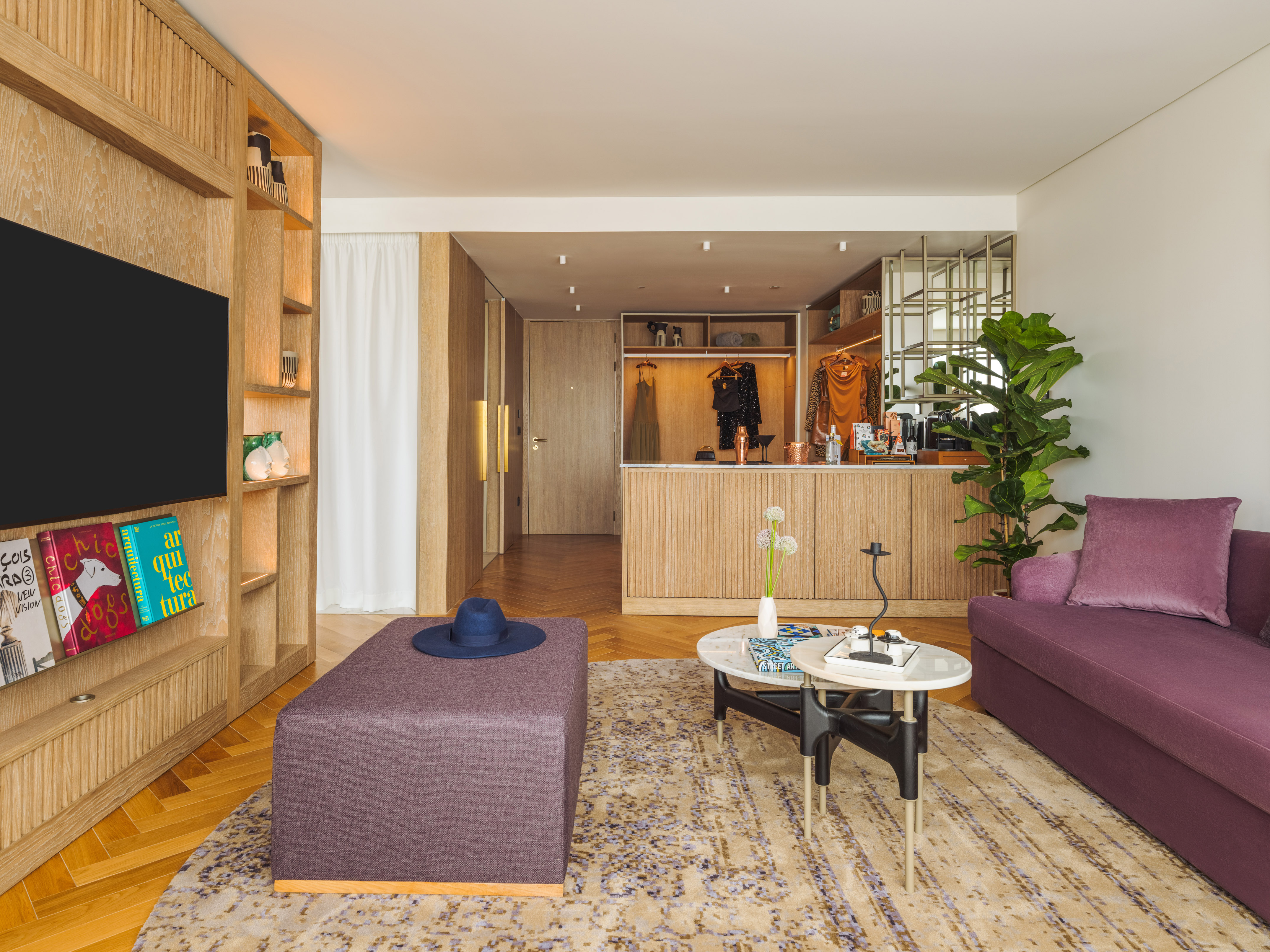 Modern hotel suite with purple sofa, wood TV unit, patterned rug, and open closet.