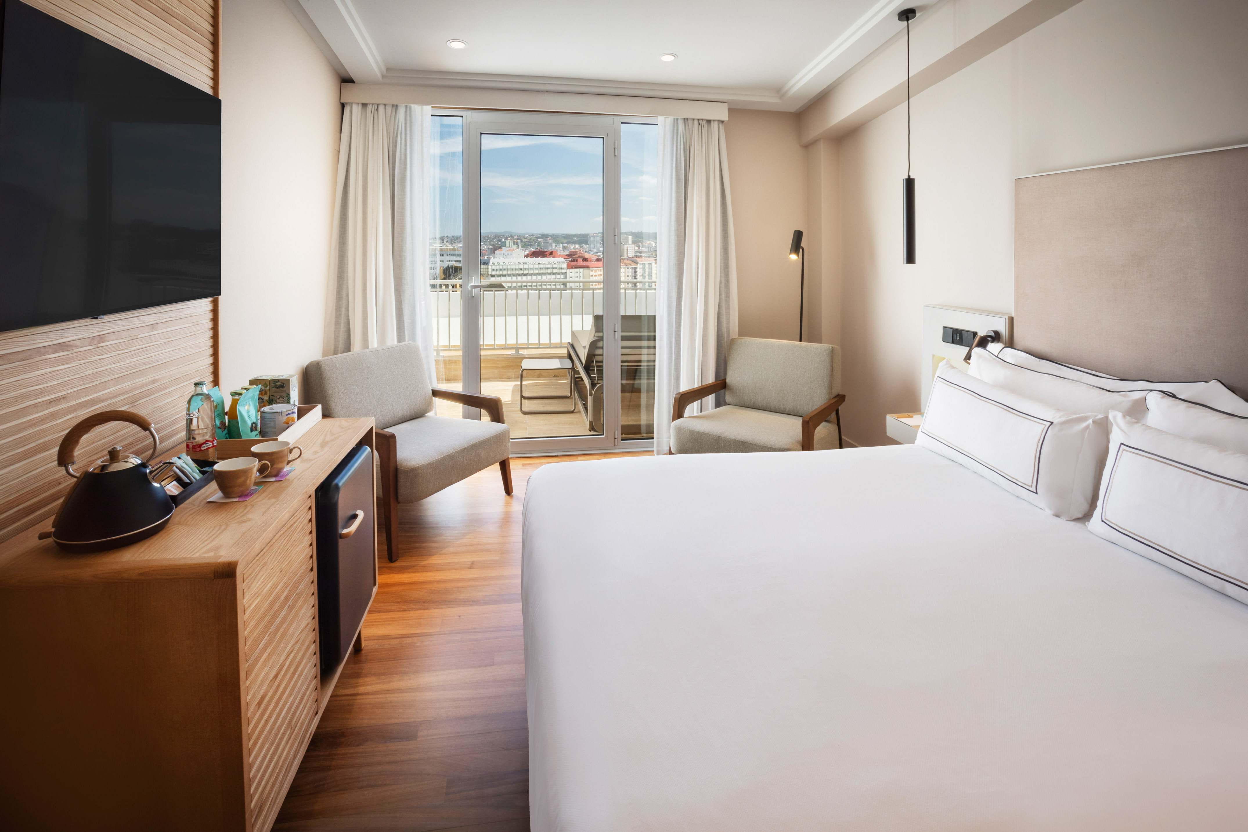 Hotel room with king bed, balcony city view, coffee station, and armchairs.