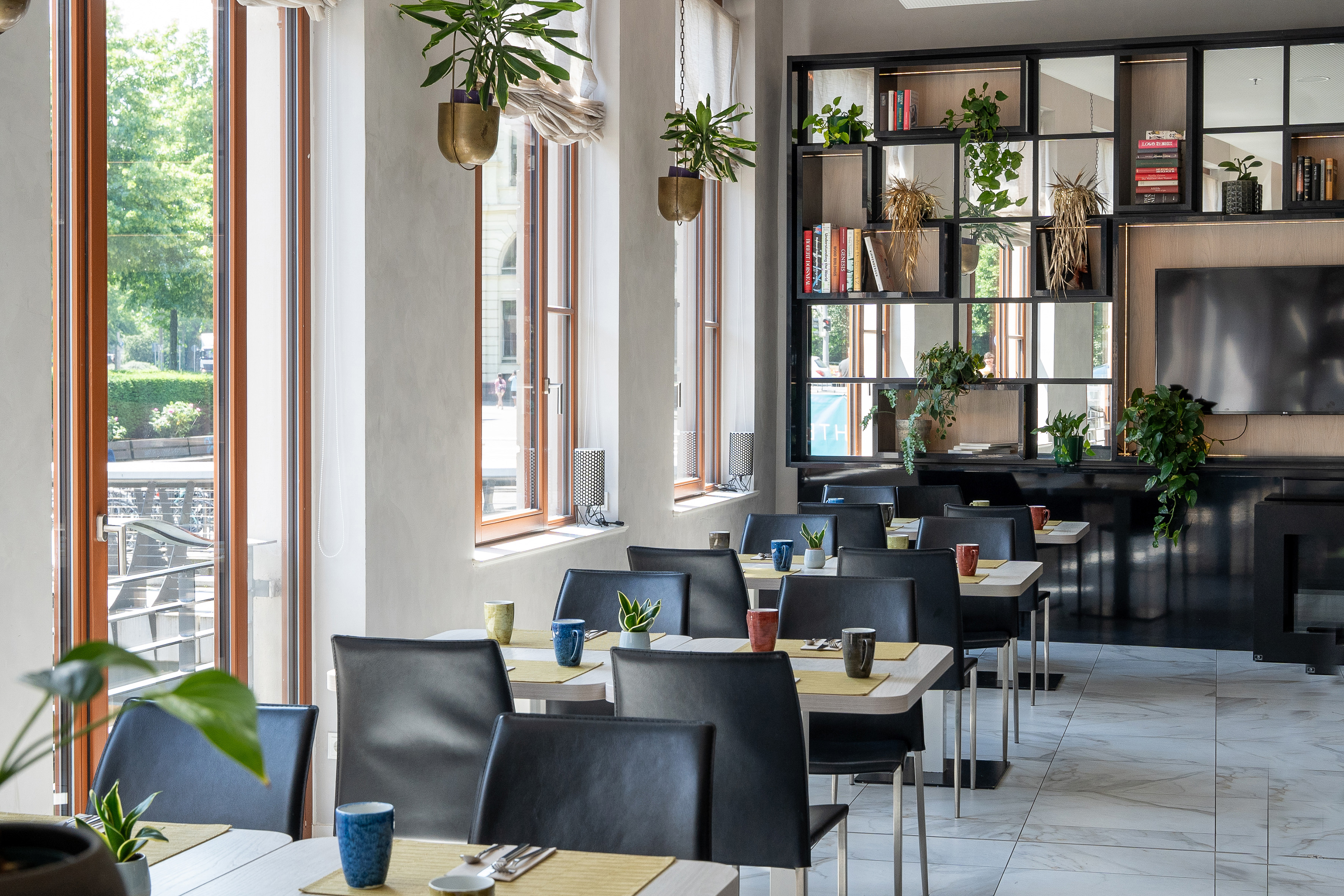 Modern restaurant dining area with tables, chairs, plants, and large windows.