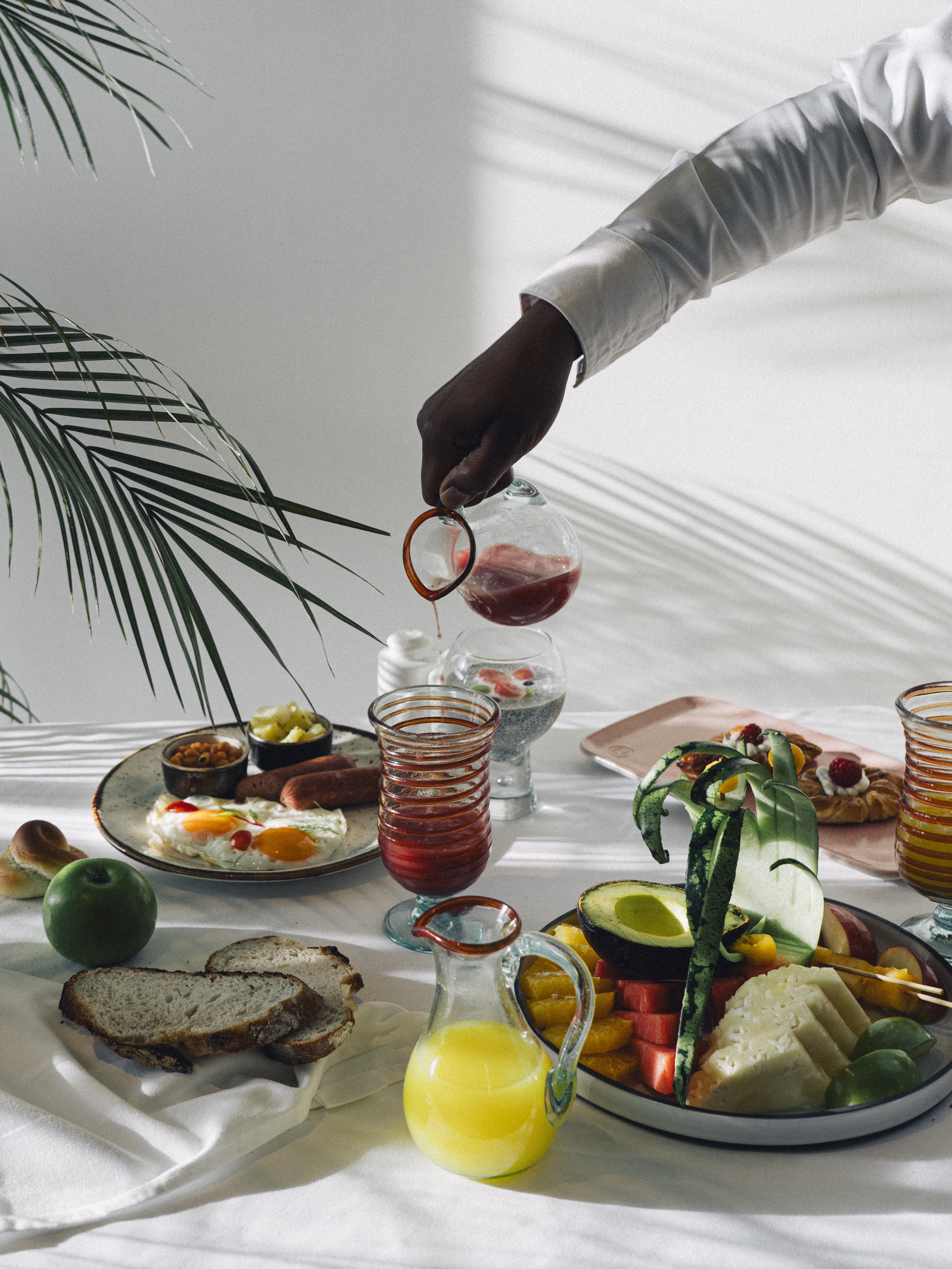 Resort breakfast: Hand pours juice over eggs, fruit platter, bread, palm shadows.