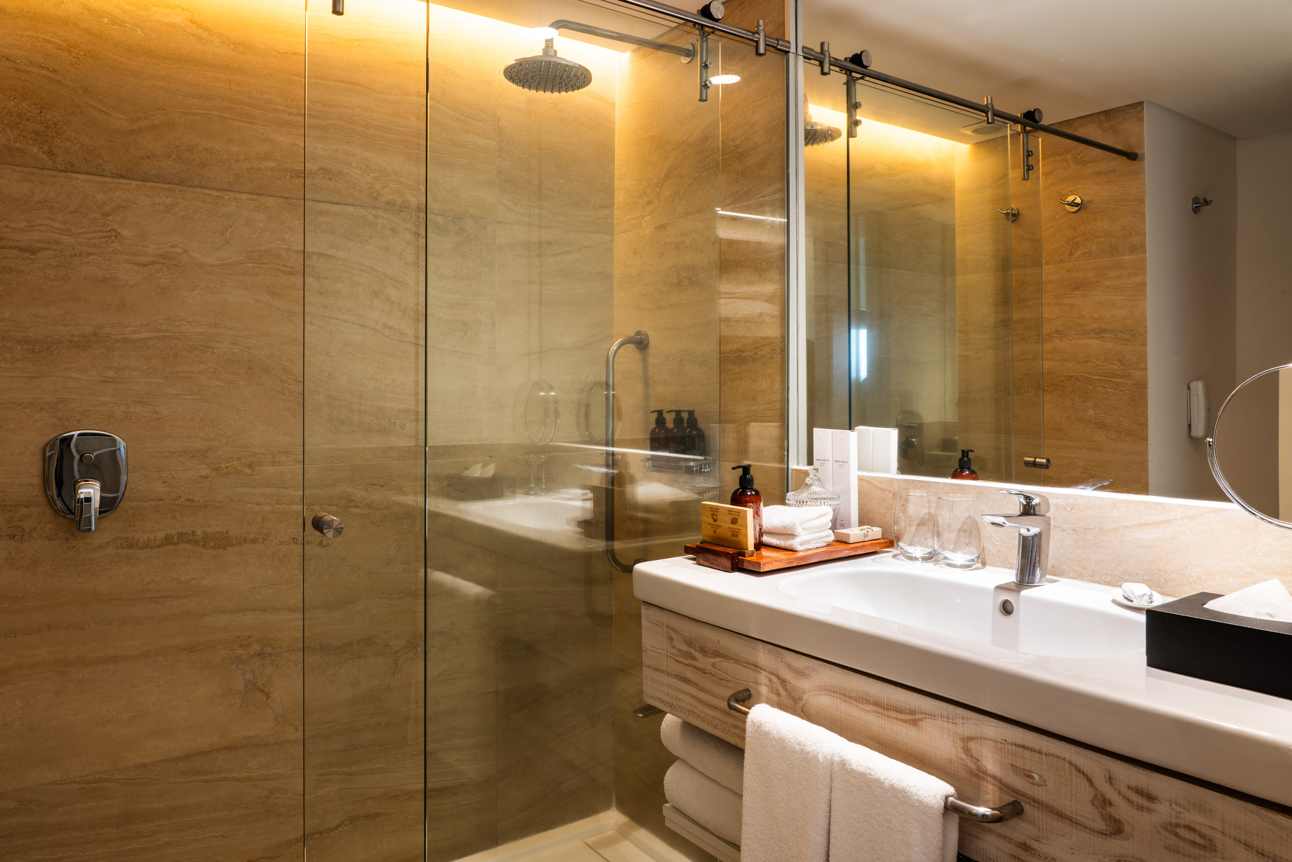 Contemporary hotel bathroom with glass shower, stone walls, and wood vanity.