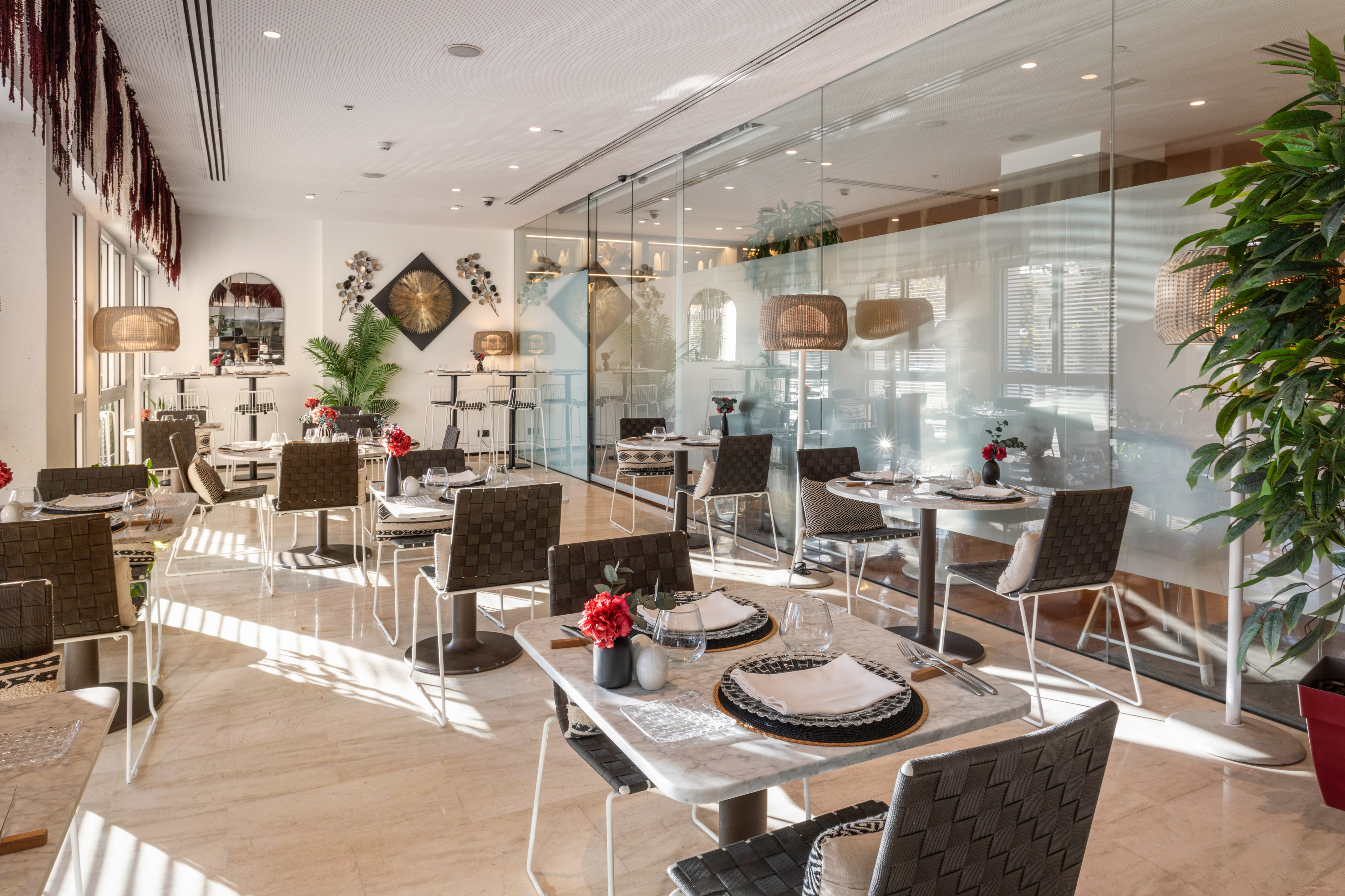 Elegant sunlit restaurant with set tables, woven chairs, and glass walls.