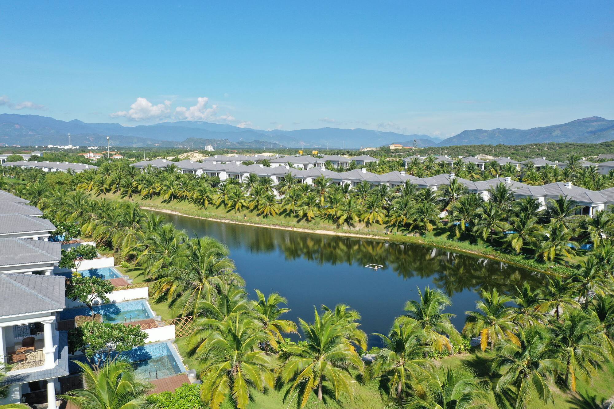 Luxury resort villas by a lake, surrounded by palm trees and mountains.