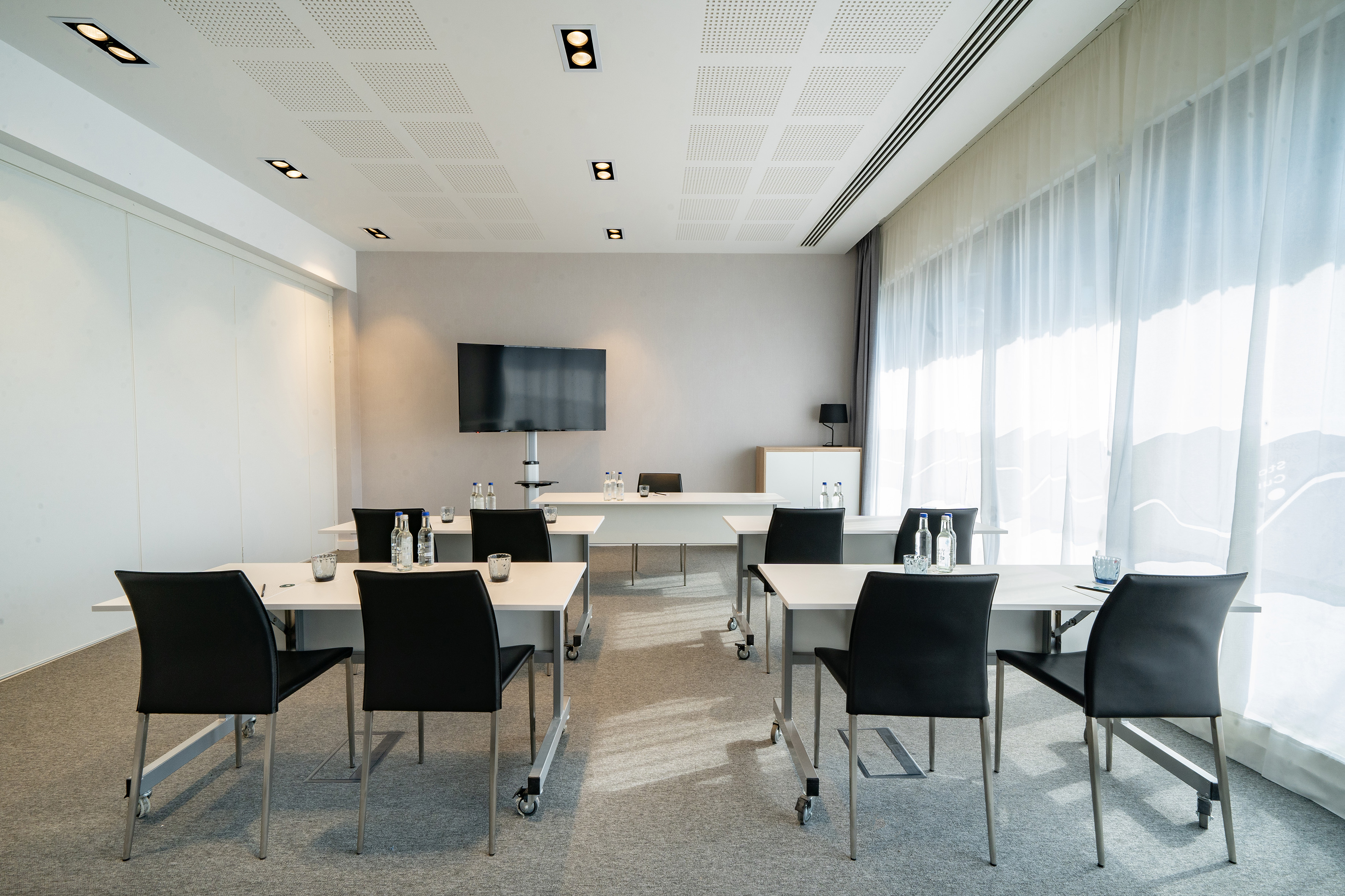 Modern conference room with tables, black chairs, large TV screen, bright windows.