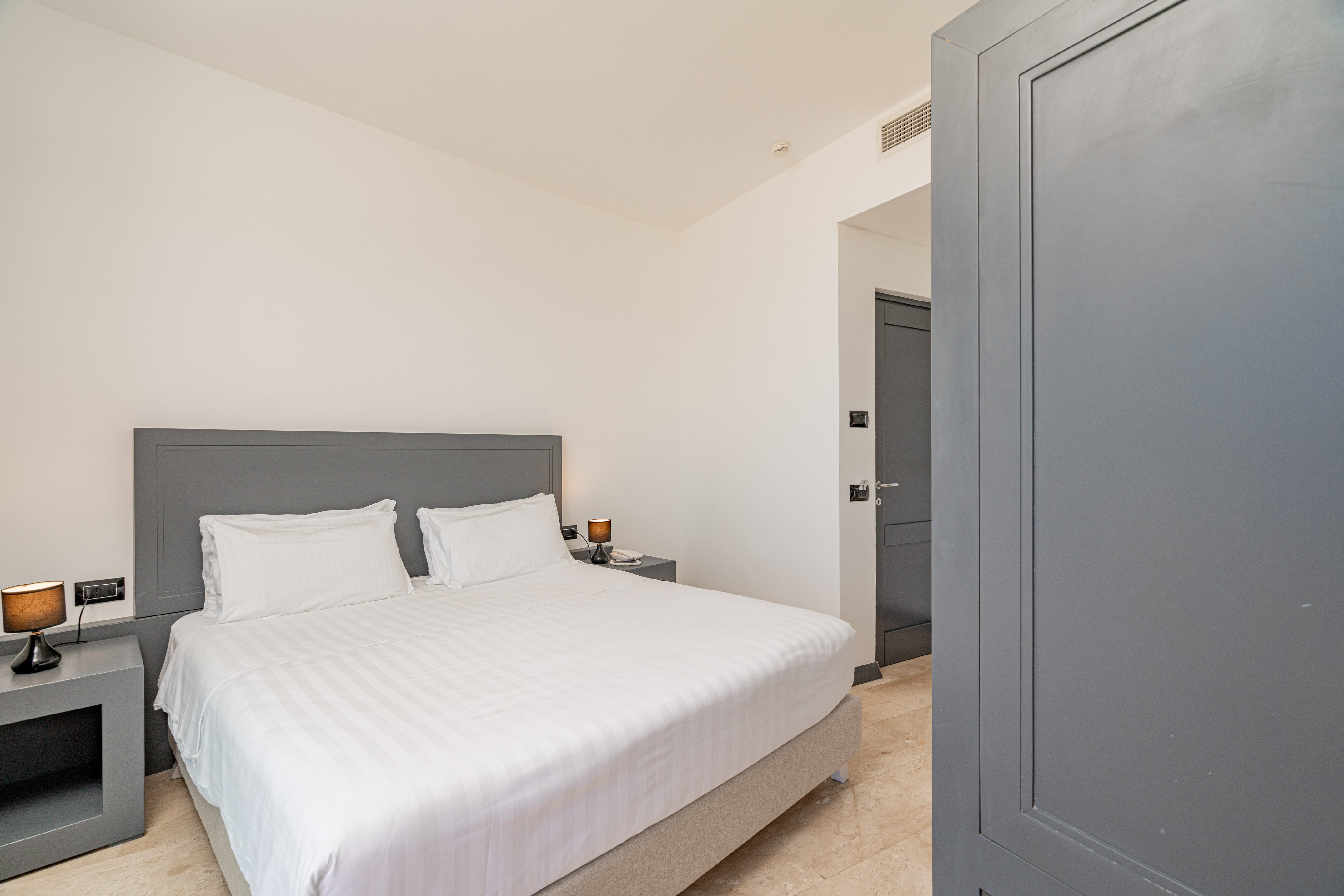 Modern hotel bedroom with grey headboard, white linen bed, and bedside lamps.
