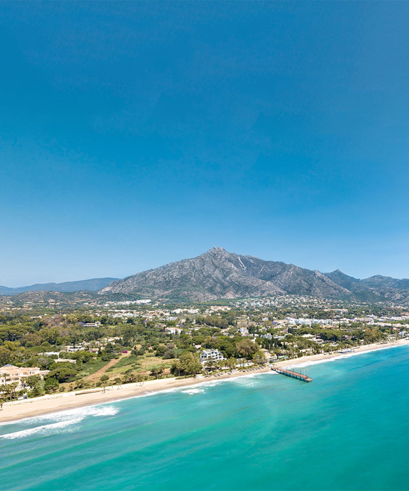 Aerial view of turquoise sea, sandy beach, coastal resort, mountains, and pier.
