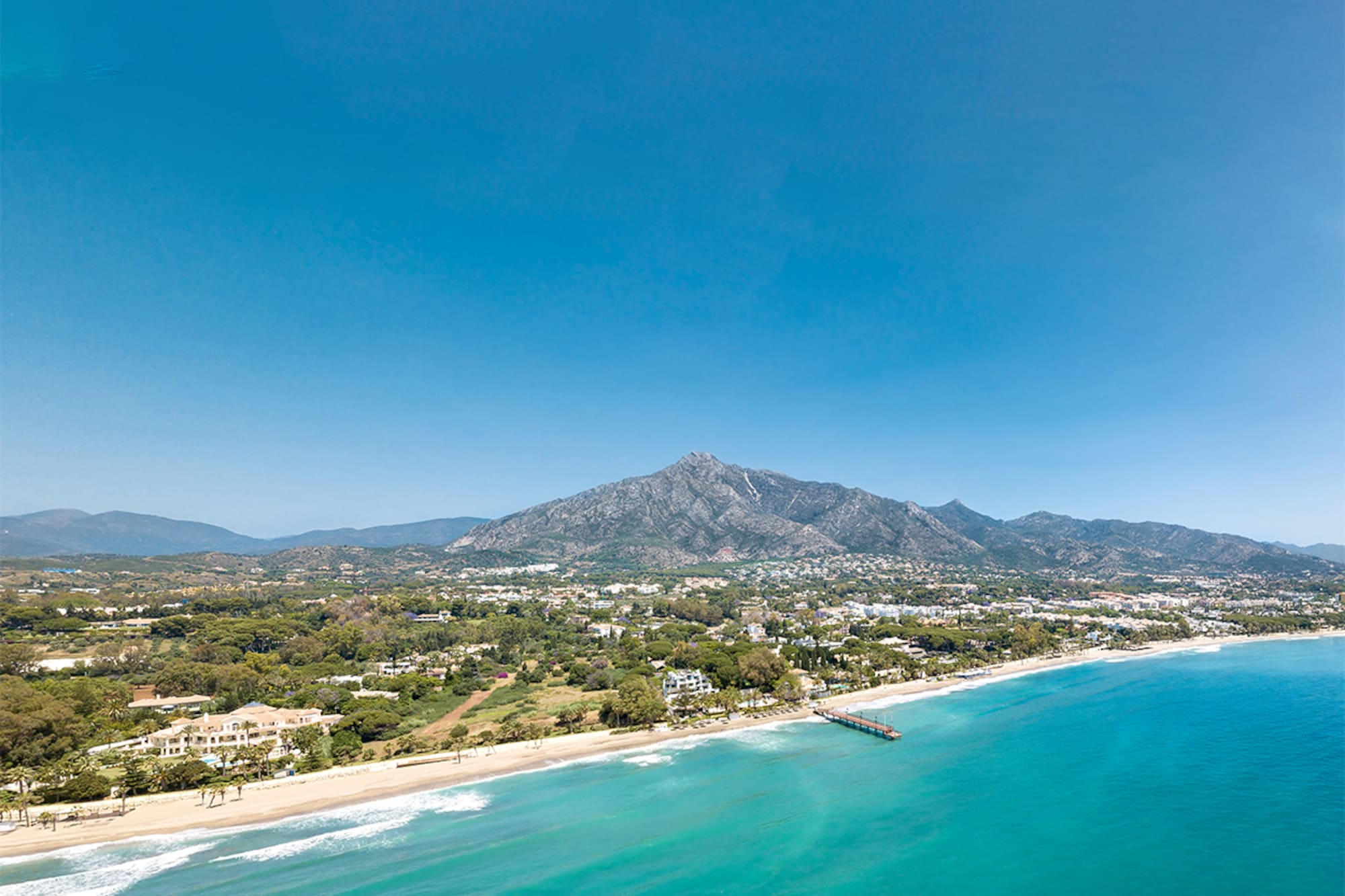 Aerial view of turquoise sea, sandy beach, coastal resort, mountains, and pier.