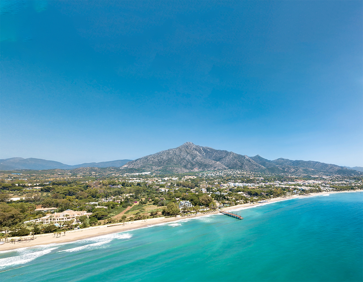 Aerial view of turquoise sea, sandy beach, coastal resort, mountains, and pier.