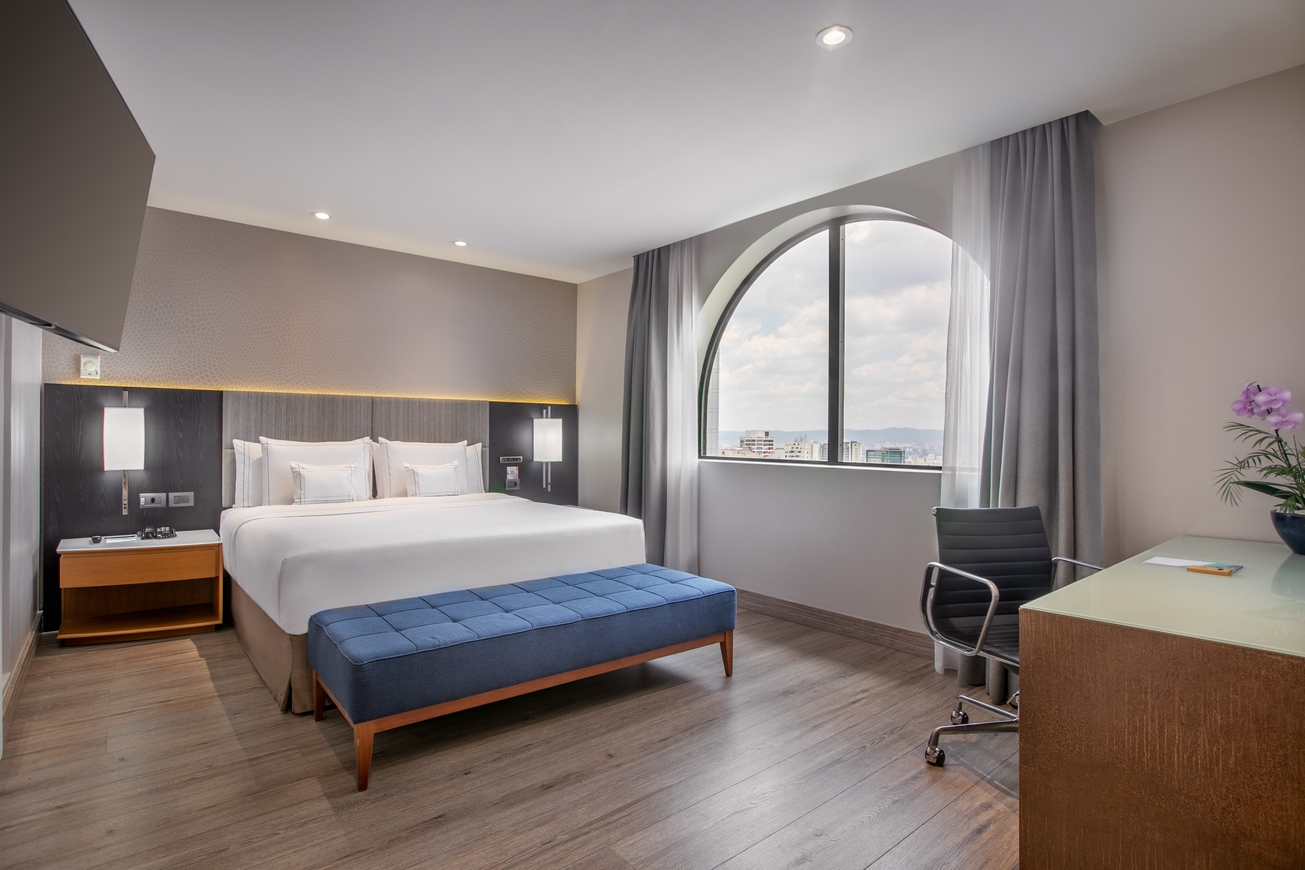 Modern hotel bedroom with king bed, desk, and city view from arched window.