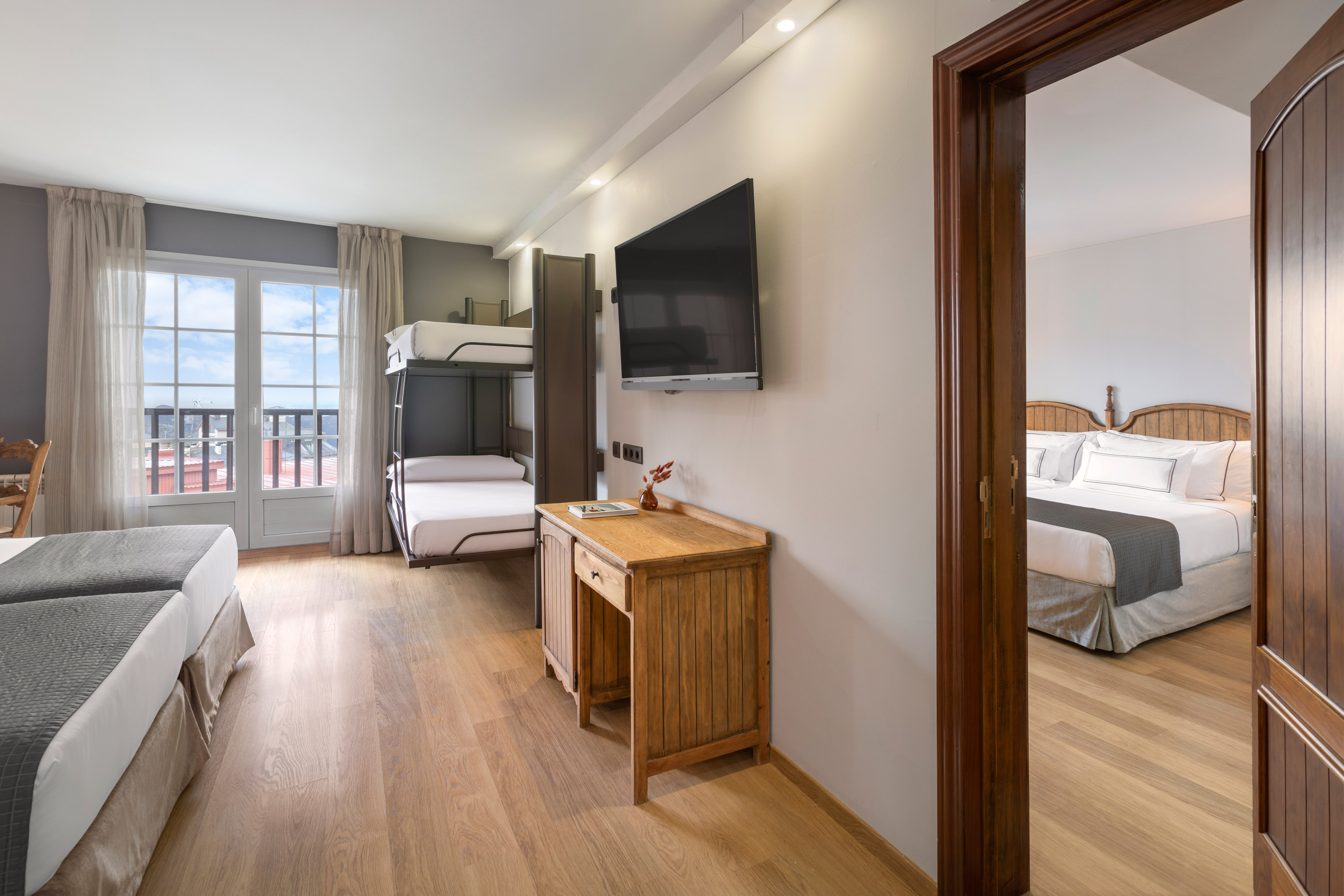 Hotel room with twin beds, bunk beds, desk, TV, window, and adjoining room.