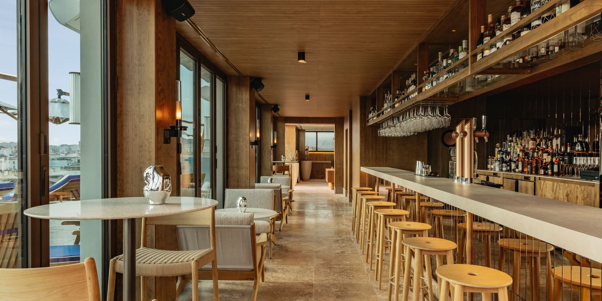 Wooden bar interior with stools, liquor, and large windows offering city views.