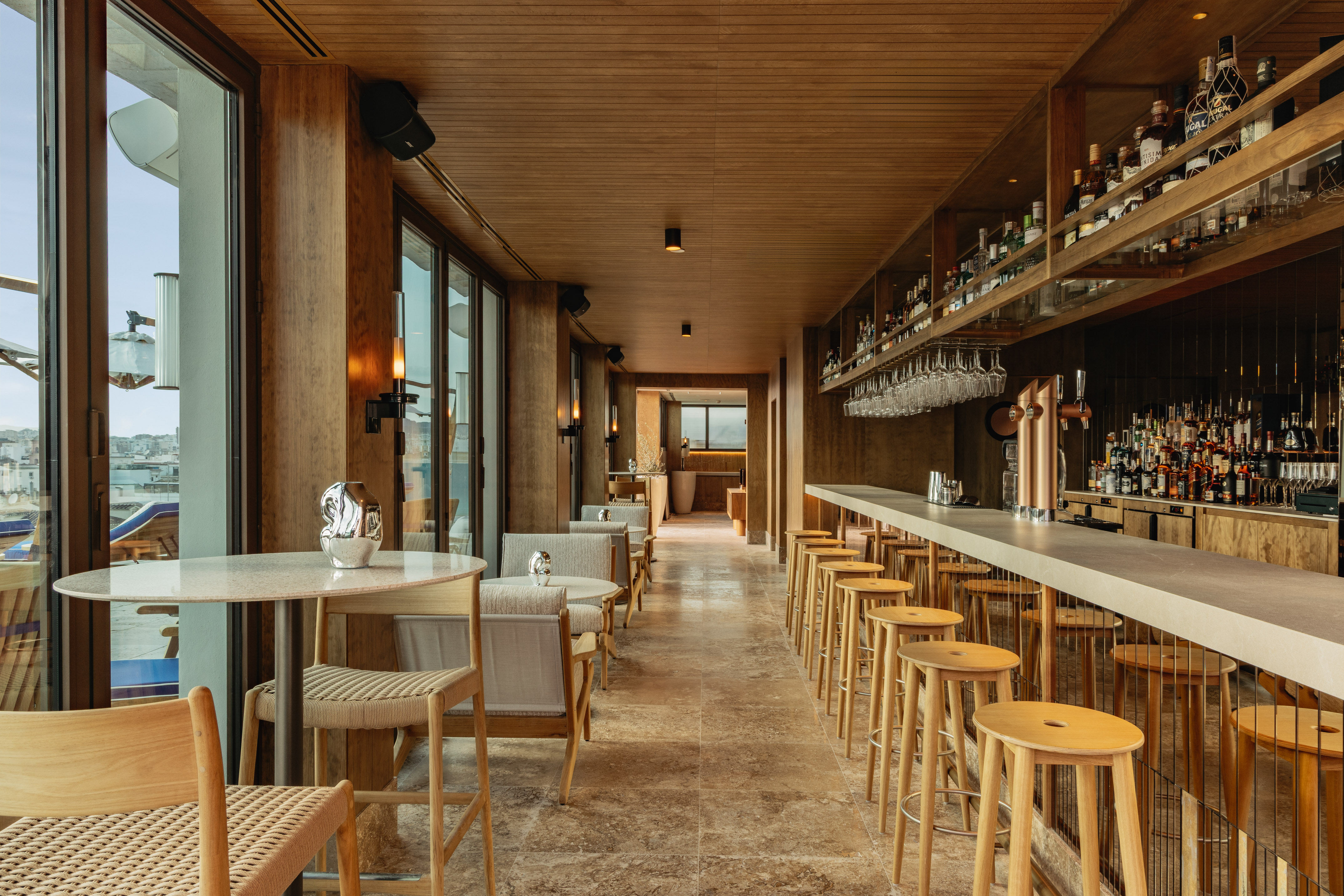 Wooden bar interior with stools, liquor, and large windows offering city views.