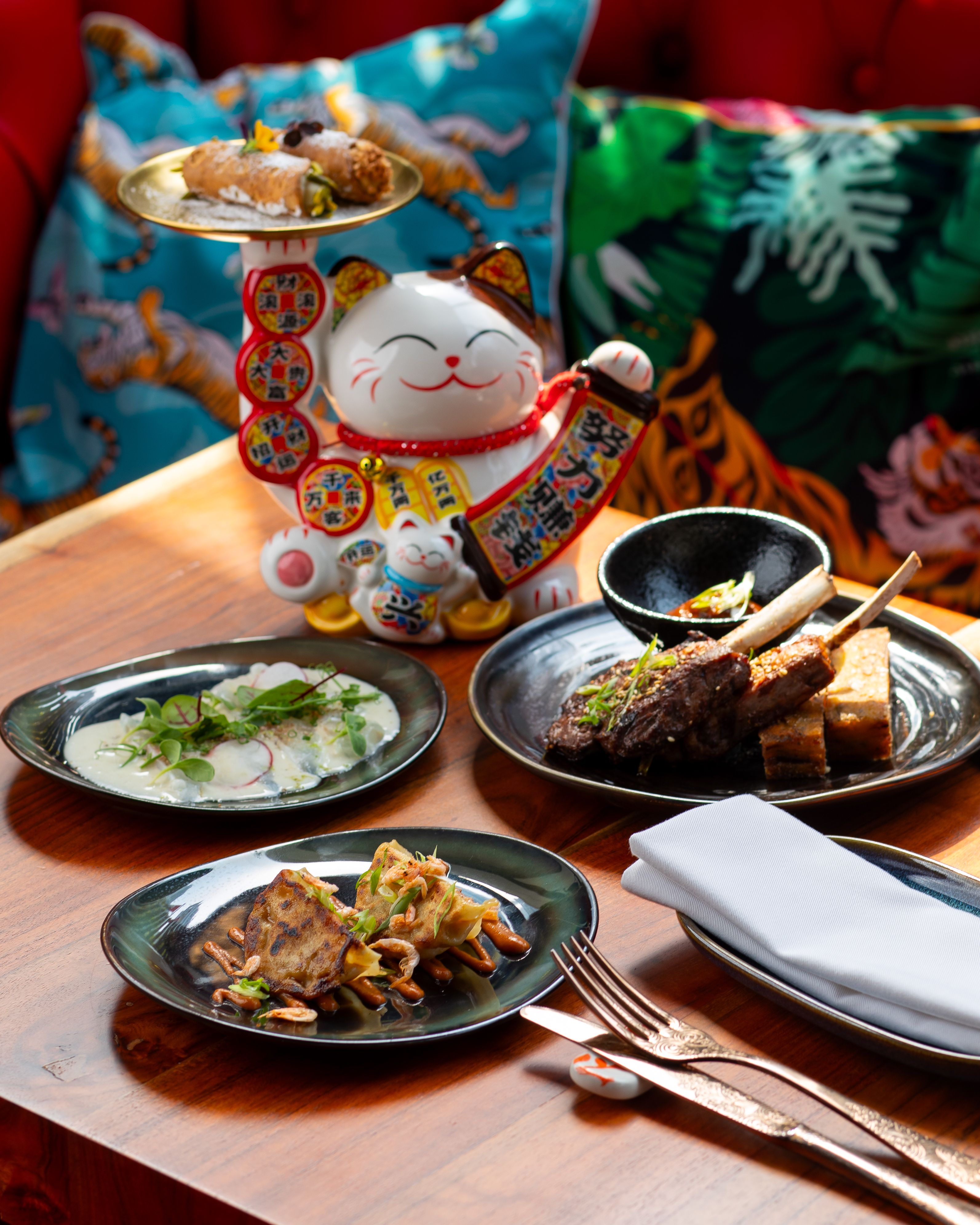 Asian fusion dishes, lamb chops, cannolis, and Maneki Neko on wooden table.