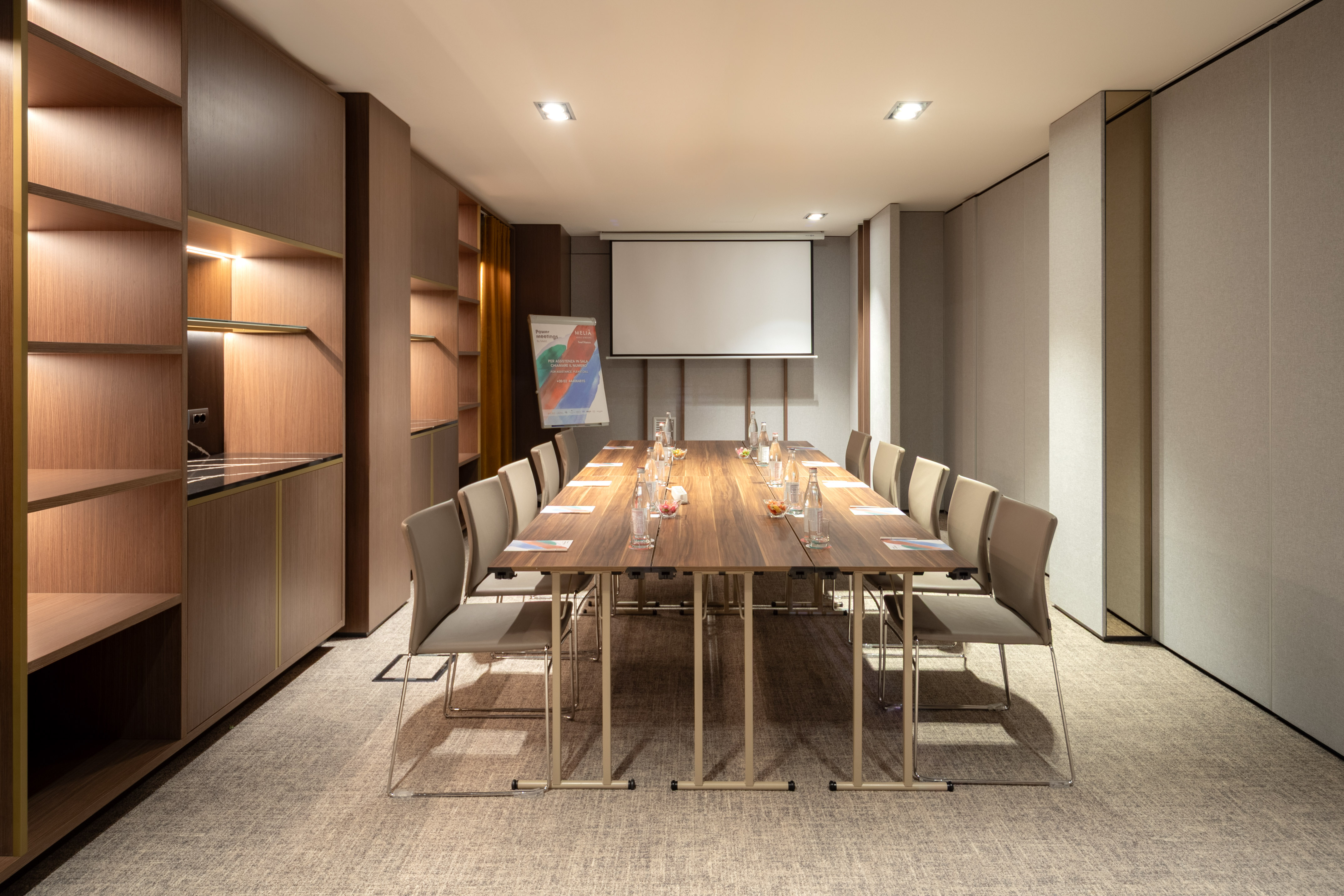 Modern conference room with wooden table, chairs, projector screen, and shelving.