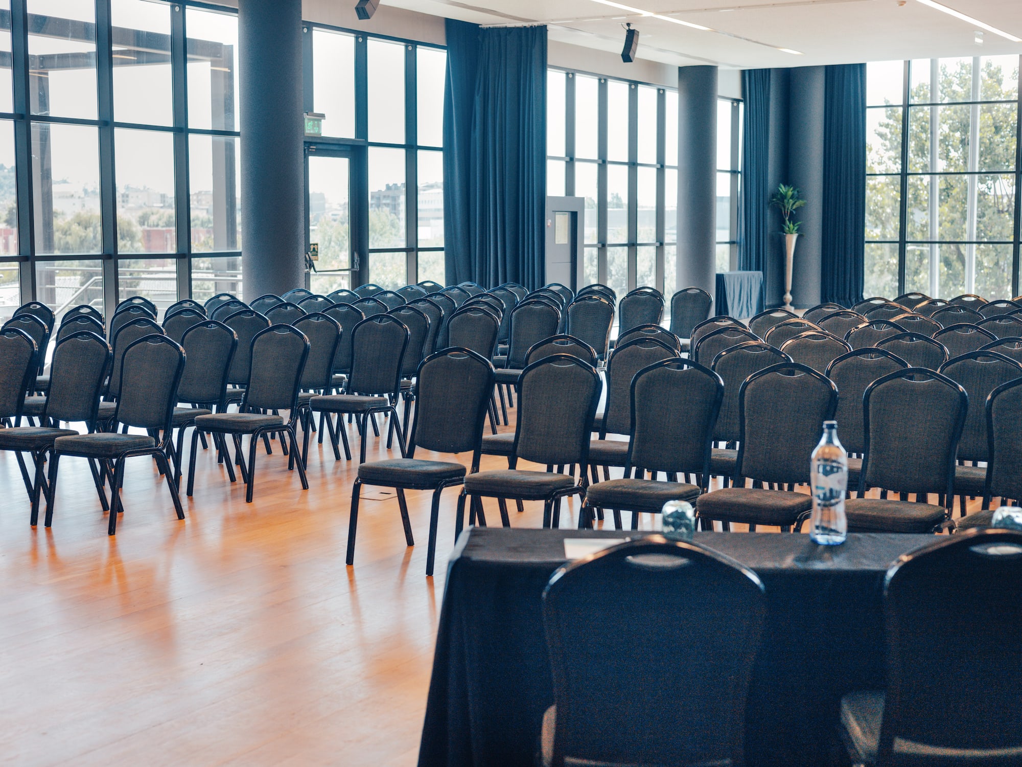 Empty conference hall with rows of dark chairs, wooden floor, and large windows.