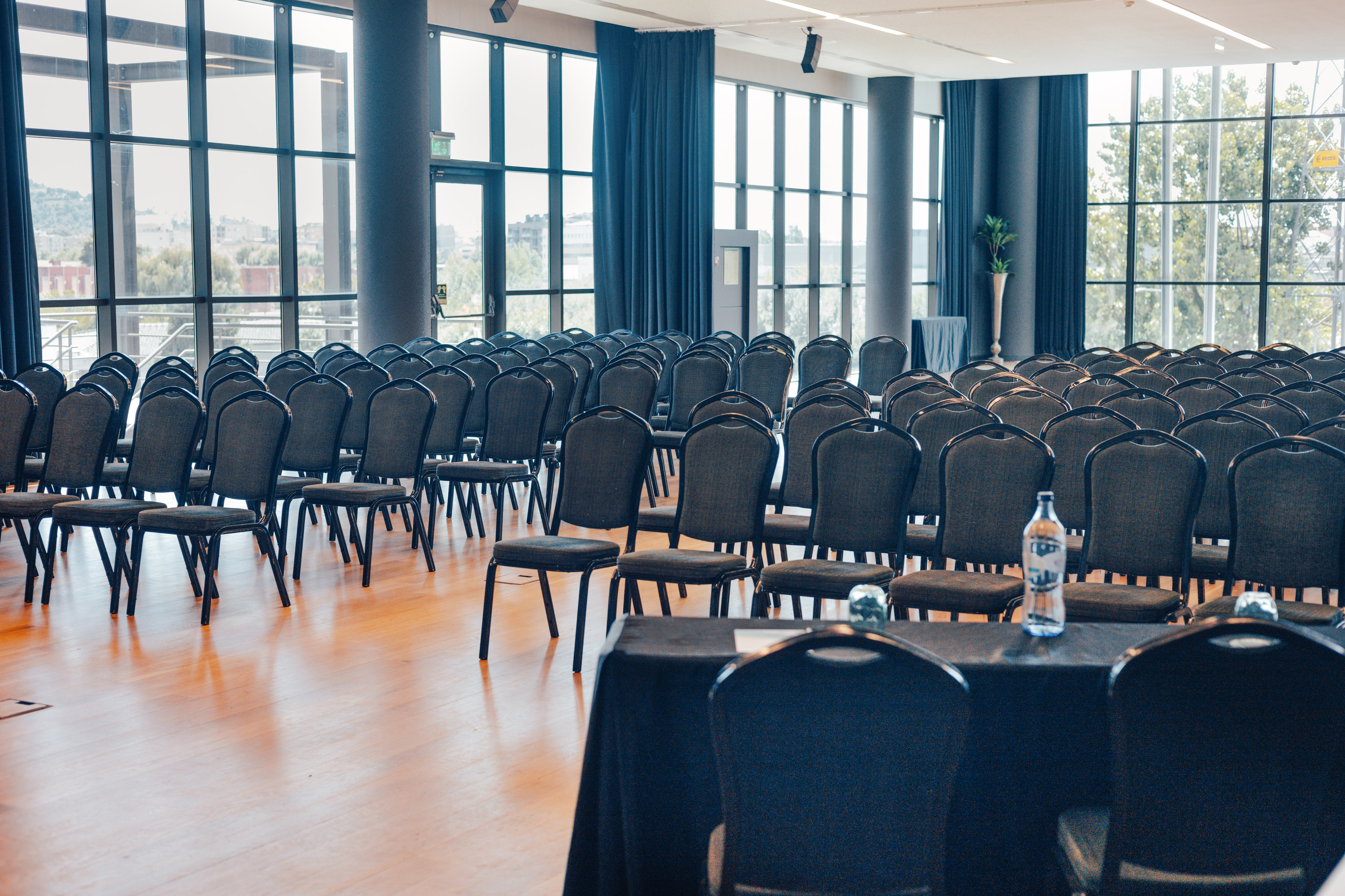 Empty conference hall with rows of dark chairs, wooden floor, and large windows.